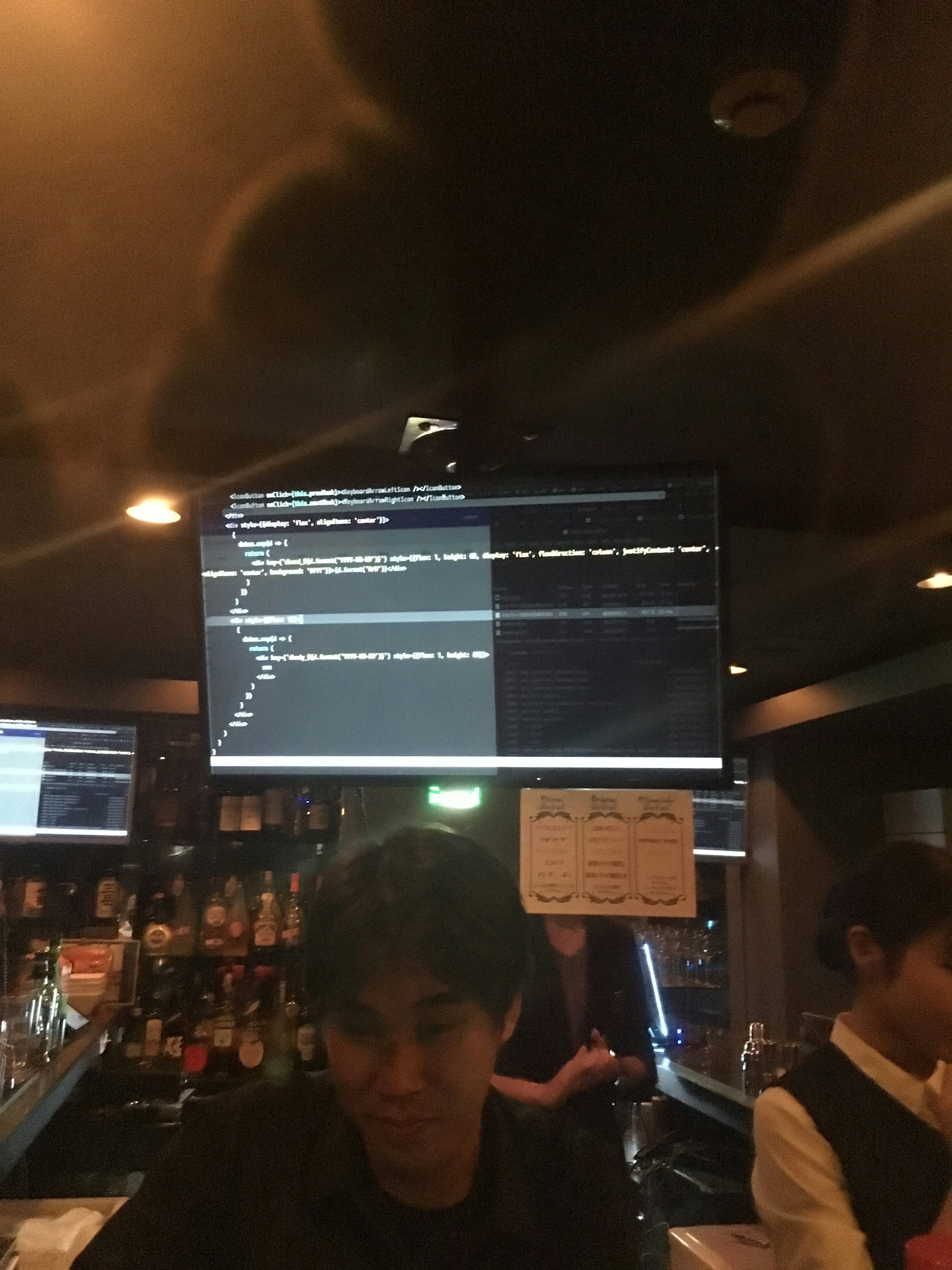 Young man working at a bar counter with a monitor displaying programming code in the background