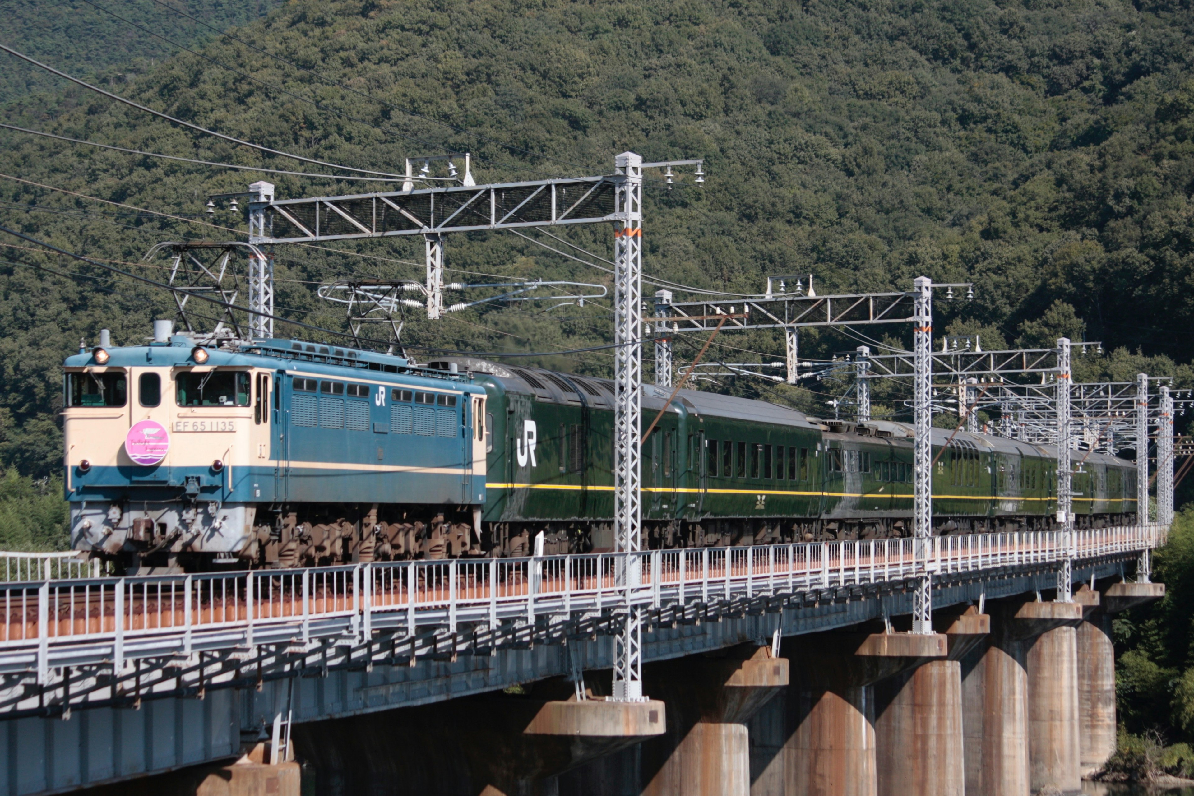 Sebuah kereta biru melintasi jembatan yang dikelilingi gunung