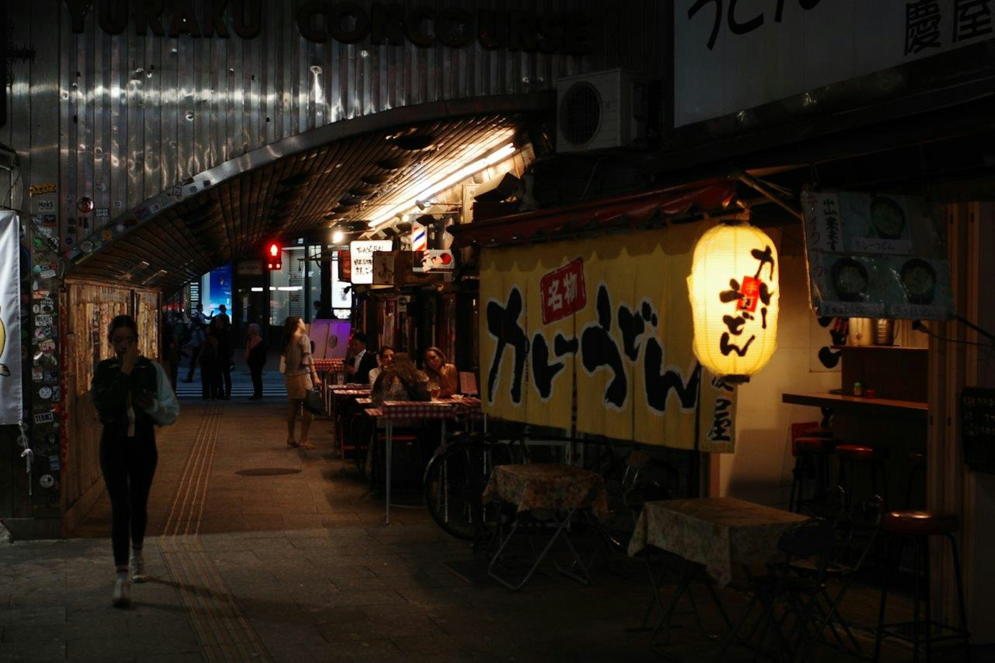 ฉากถนนที่มีป้ายร้านอาหารสว่างและคนเดินเดี่ยว