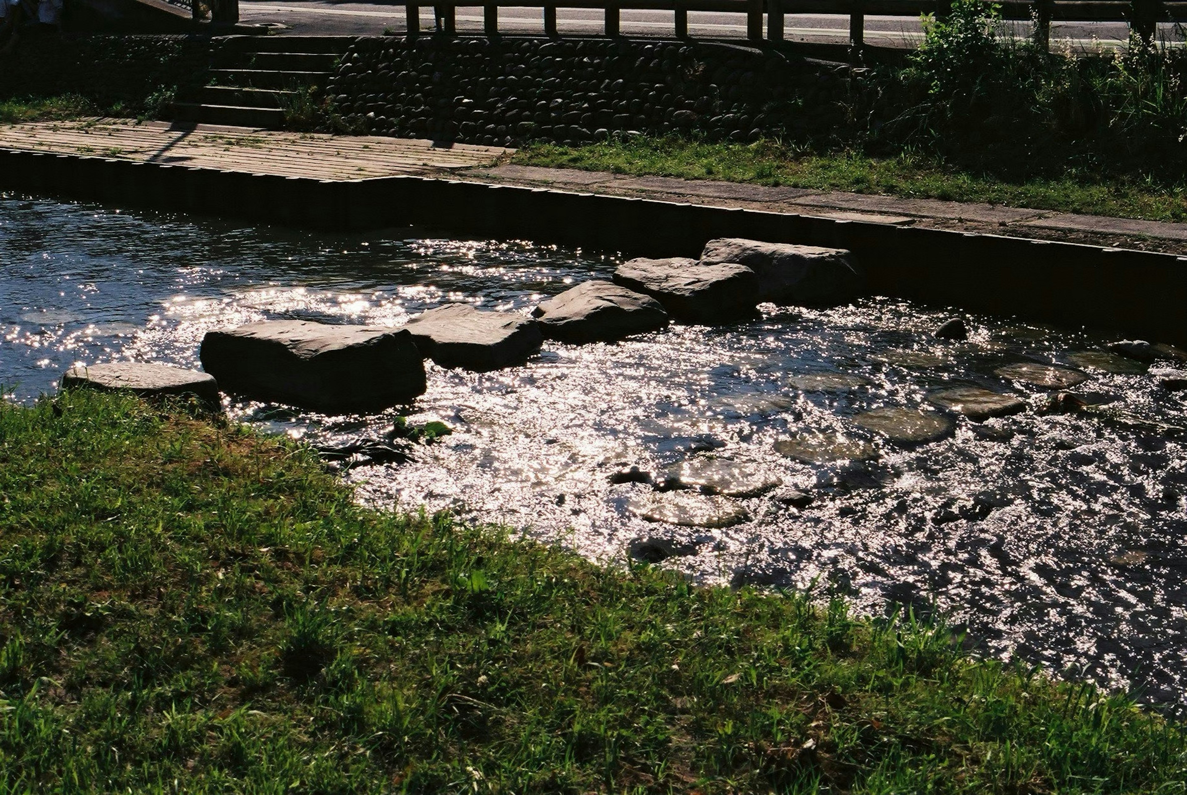 石のステップがある小川の風景 日差しを反射する水面 緑の草が生えた岸