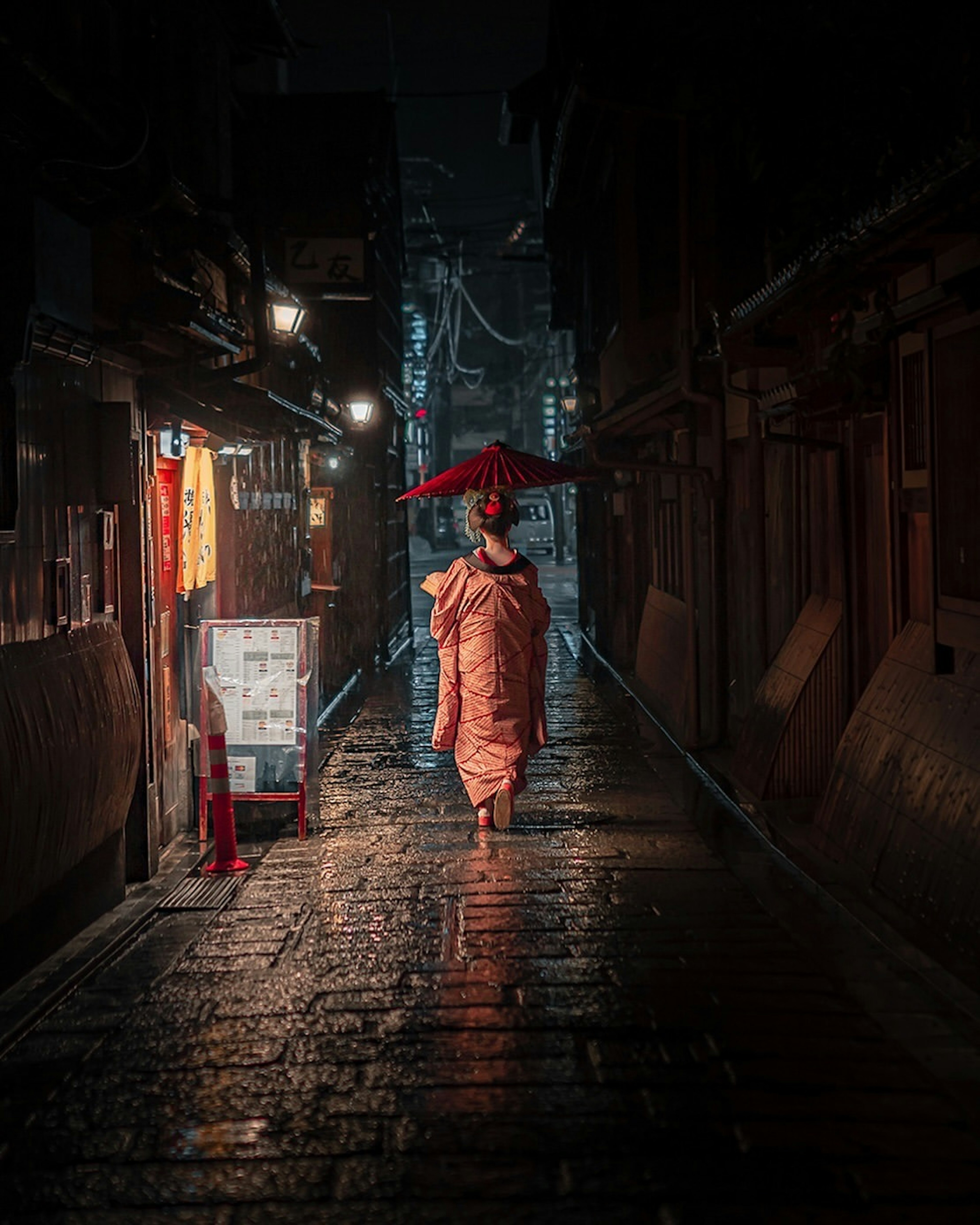 Una mujer con kimono caminando por una calle estrecha de noche con un paraguas rojo