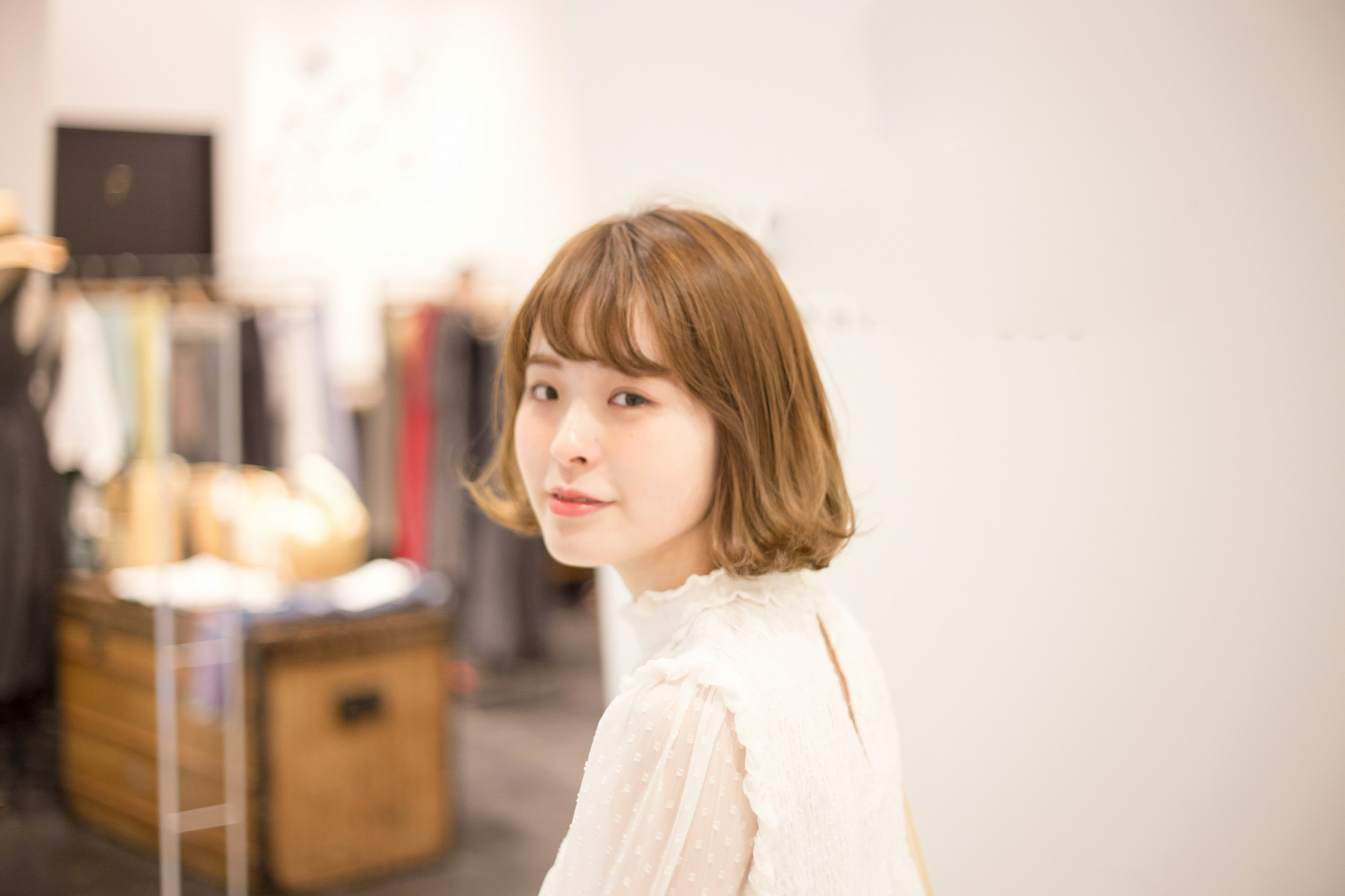 A woman looking back with a soft expression featuring short brown hair and a white outfit background includes clothing racks