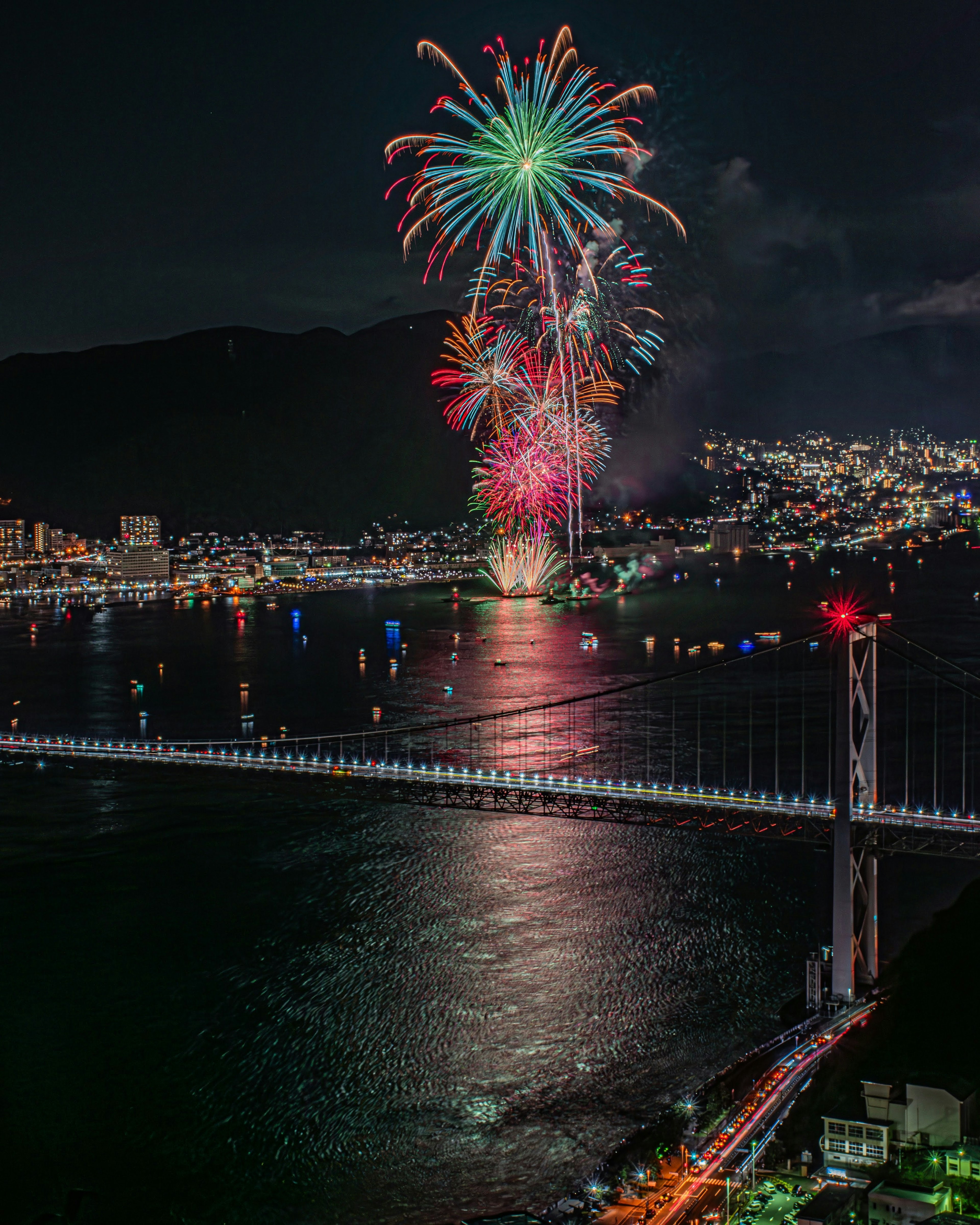 Des feux d'artifice colorés illuminent le ciel nocturne au-dessus d'une mer réfléchissante et d'un pont