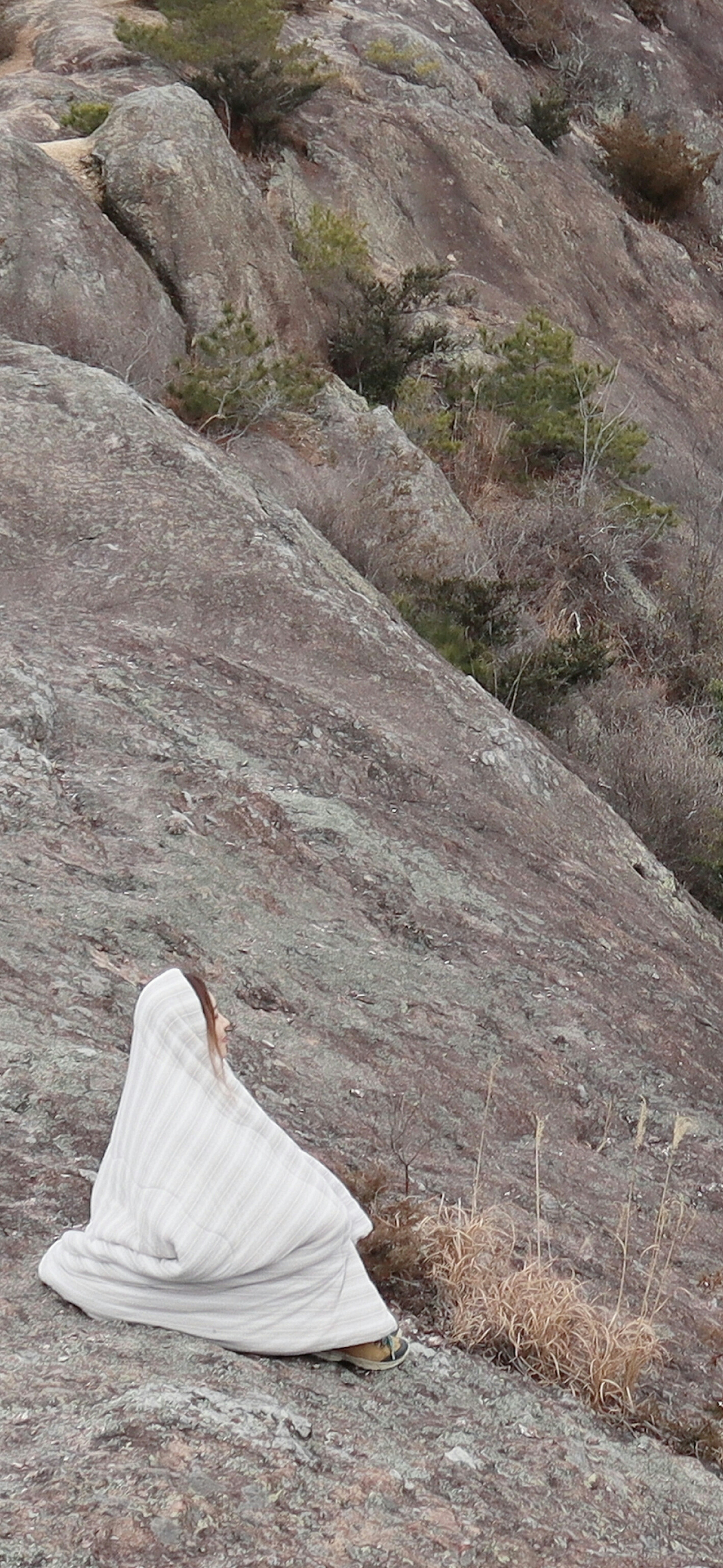 A woman draped in a white cloth stands on a rocky slope