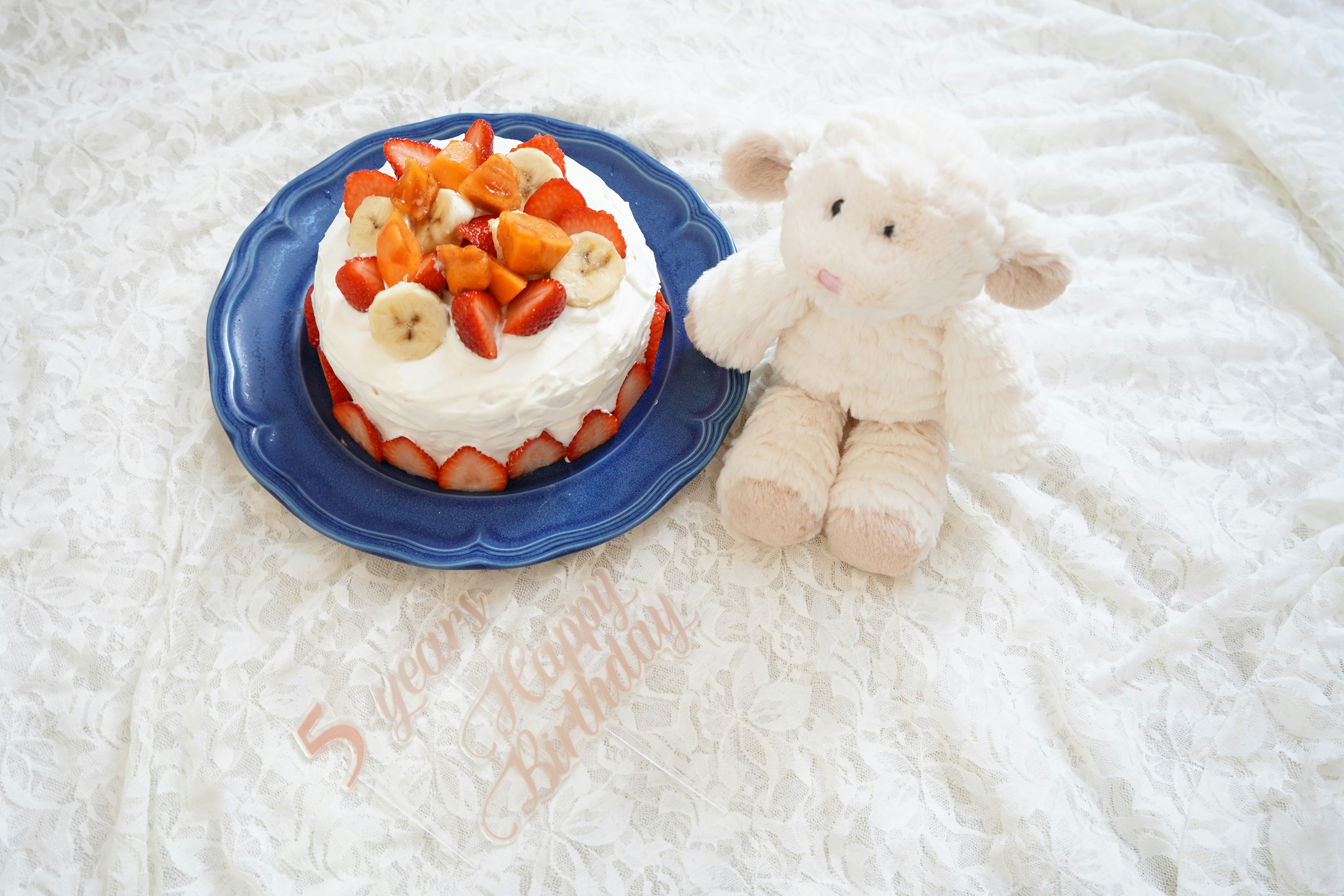 Un gâteau garni de fraises et de bananes sur une assiette bleue à côté d'un mignon mouton en peluche sur un fond blanc