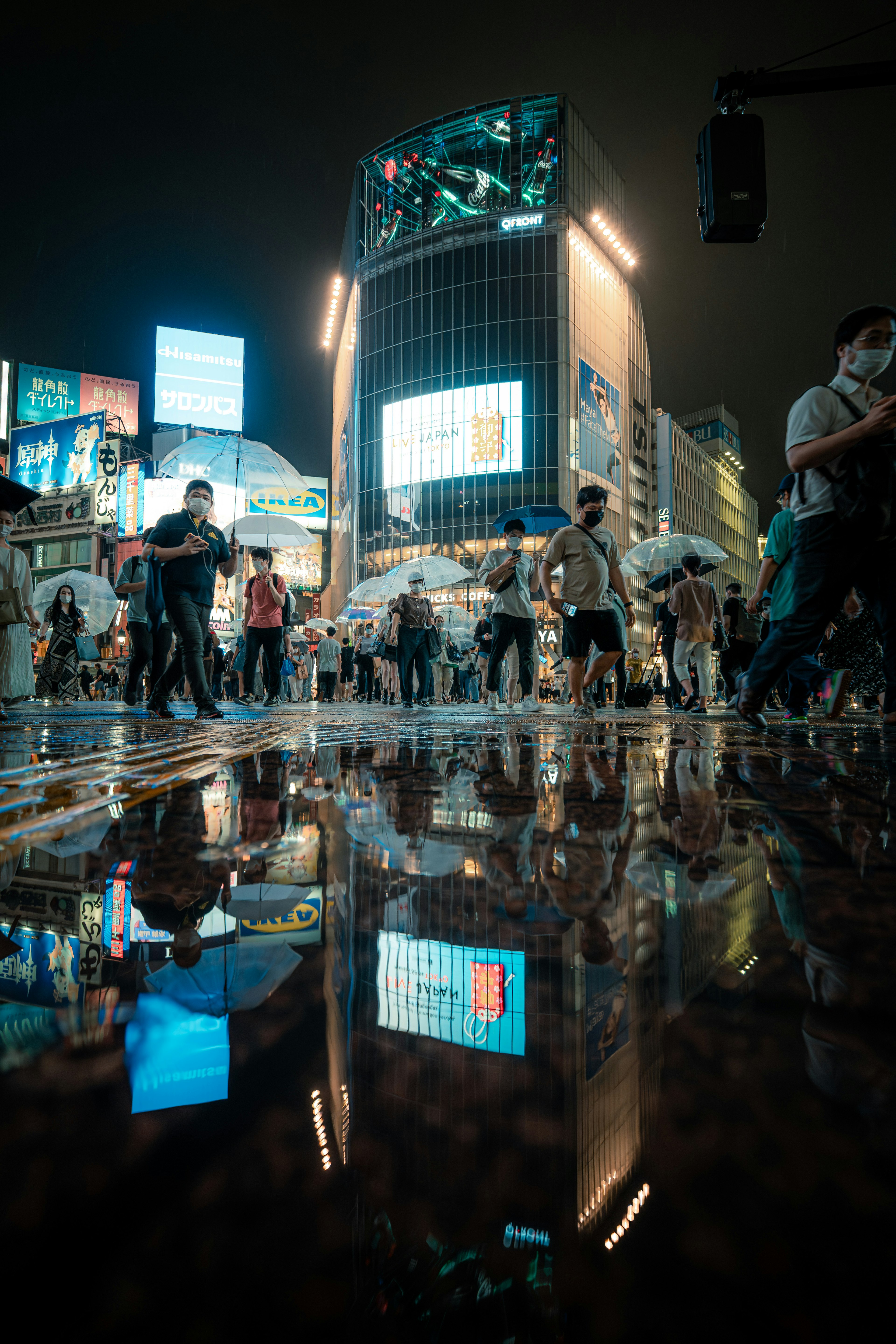 夜の渋谷の交差点で人々が行き交う様子と水たまりに映る光