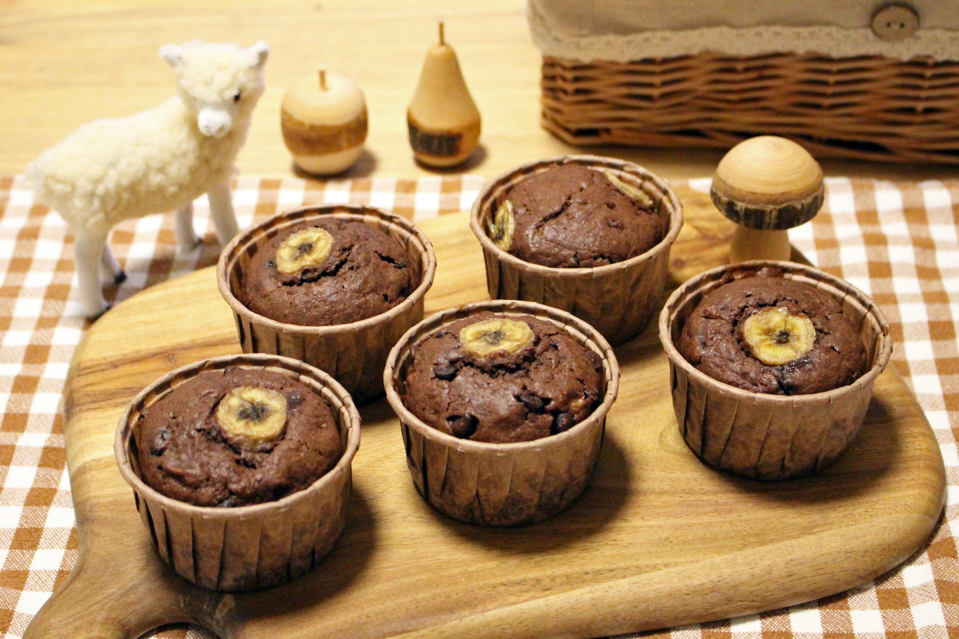 Five banana chocolate muffins arranged on a wooden board with a white sheep figurine and wooden objects in the background