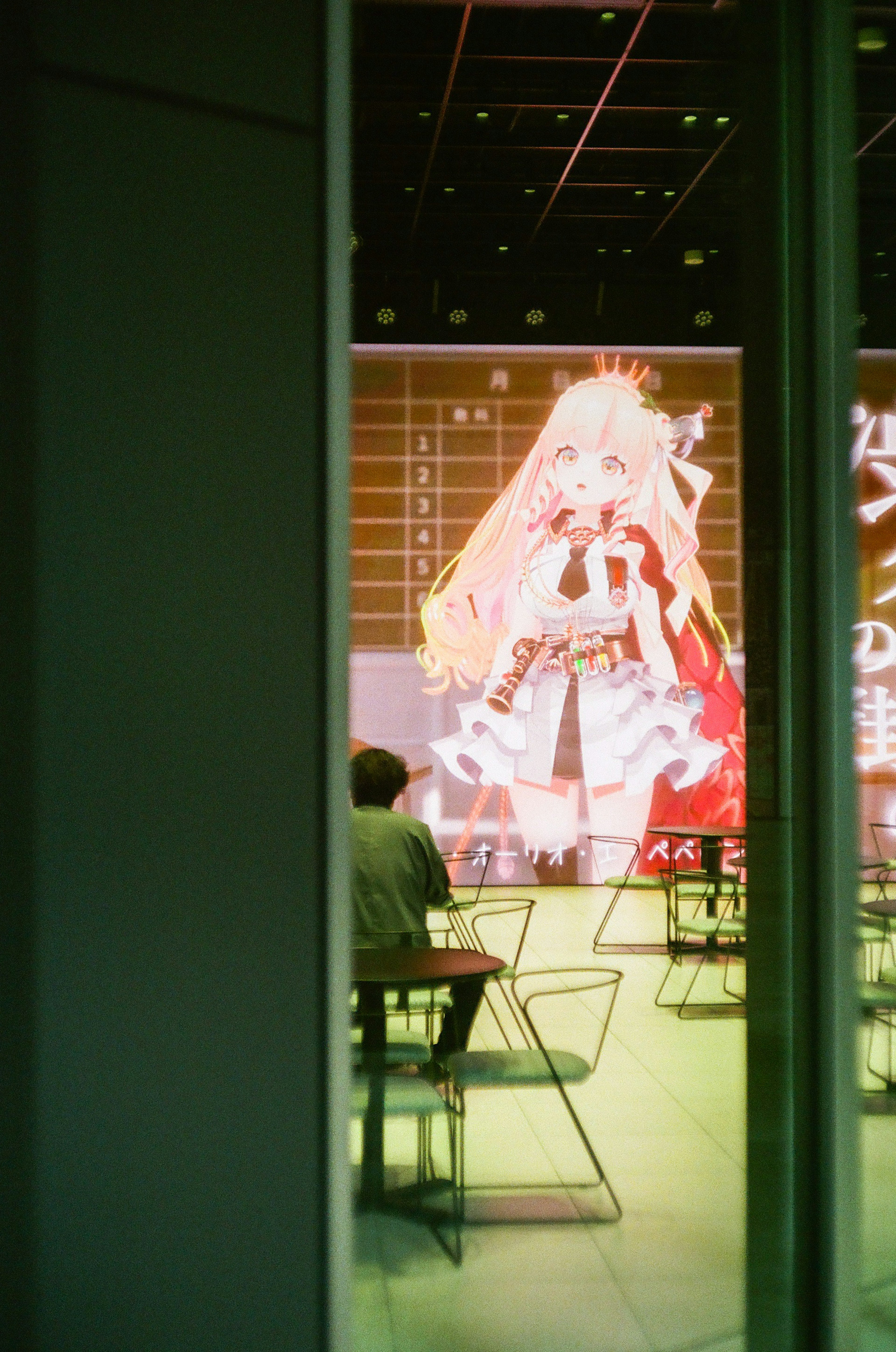 A person sitting in a café with a large anime character displayed on the wall