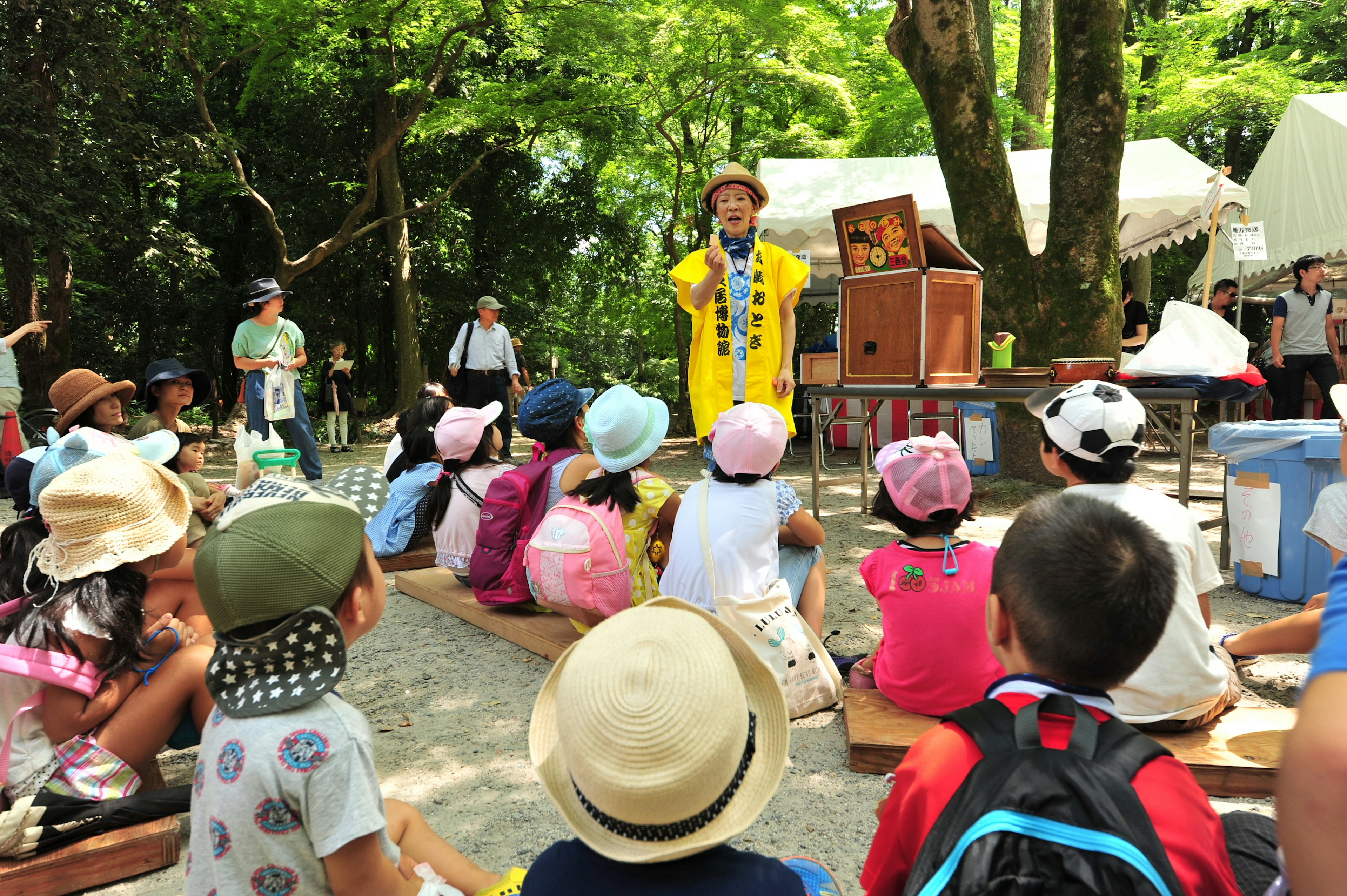 Niños disfrutando de una actuación al aire libre un artista con atuendo amarillo en el centro rodeado de niños sentados