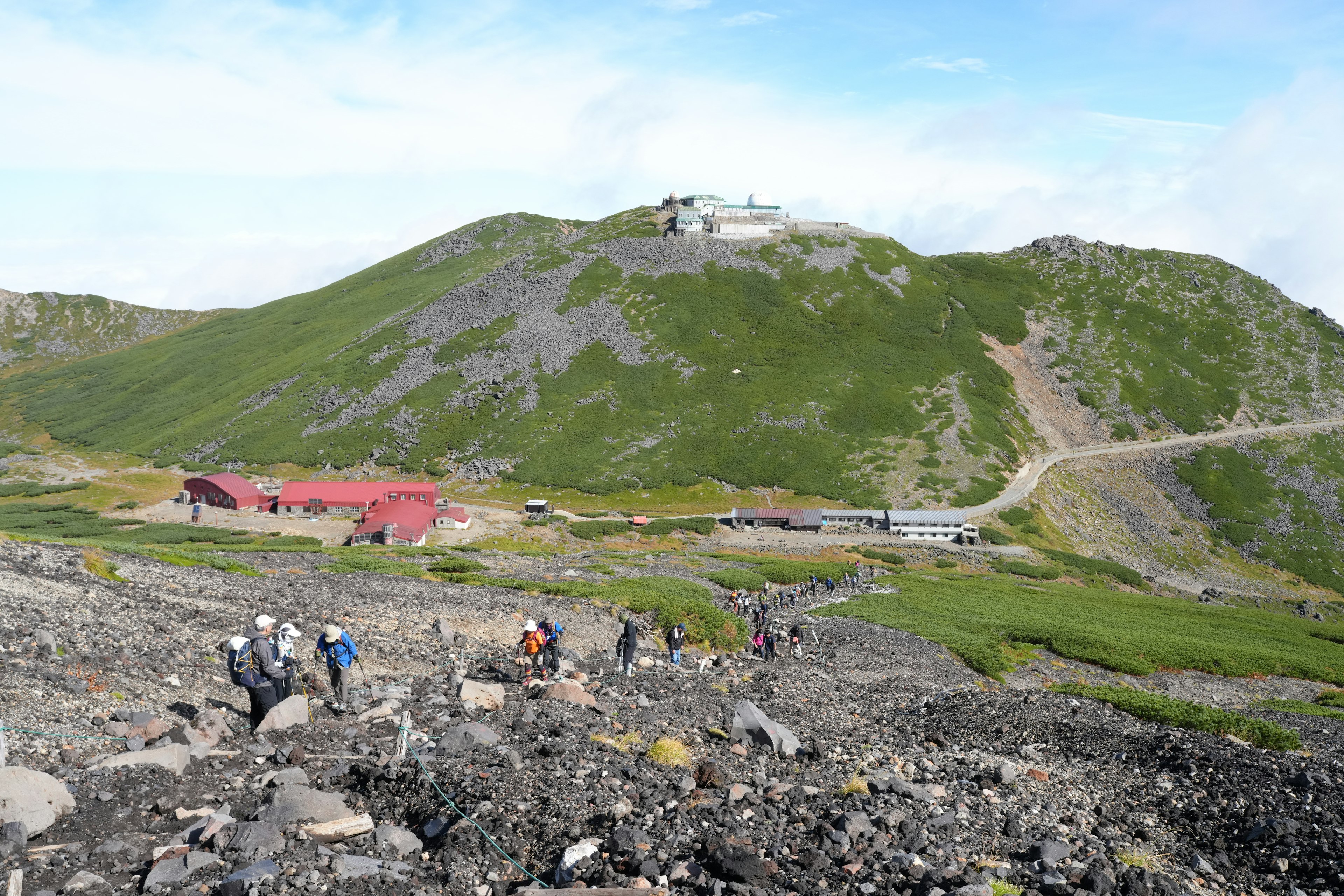 緑の山と赤い建物が見える登山道を歩く人々