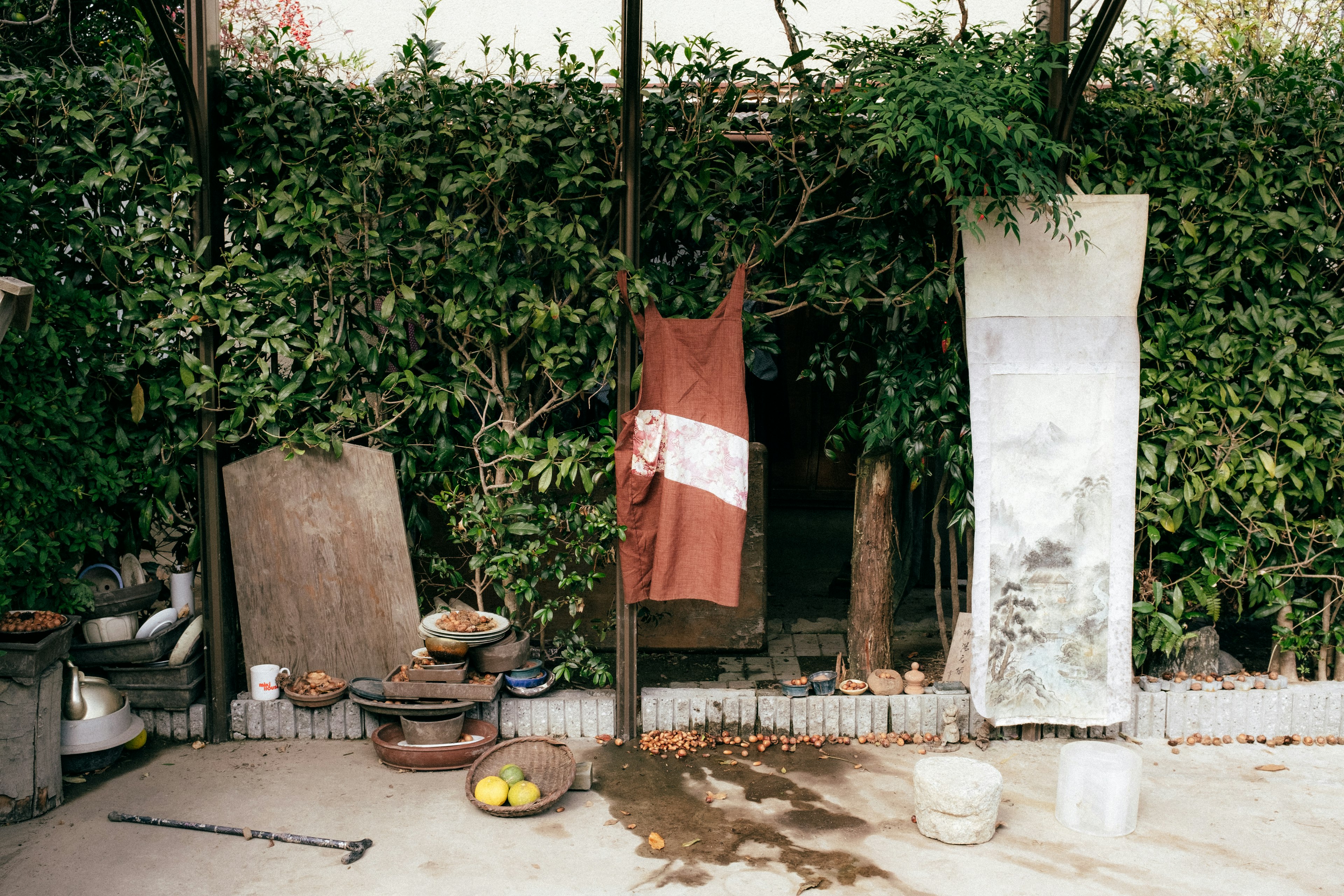 Brown apron hanging in front of a green hedge with various old tools and pots