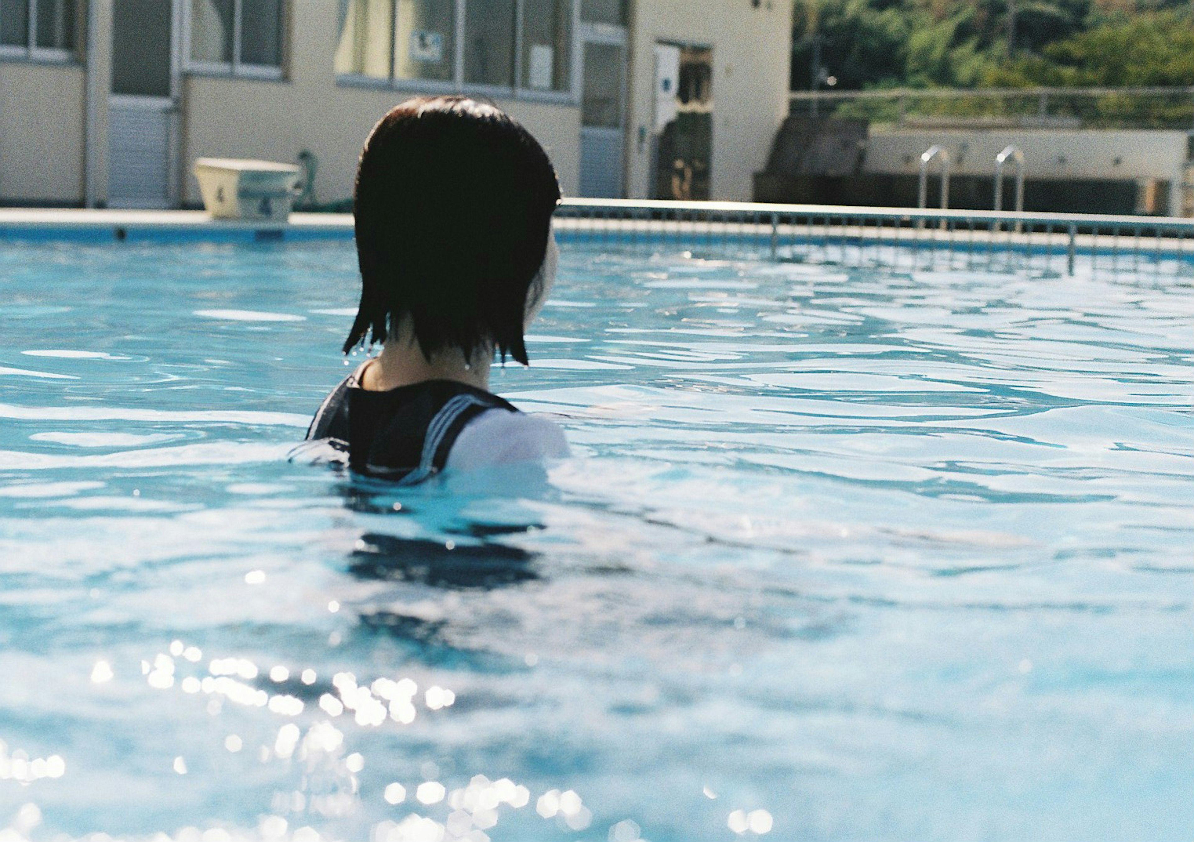 Woman seen from behind in a pool Water surface reflecting light Nearby building and natural background