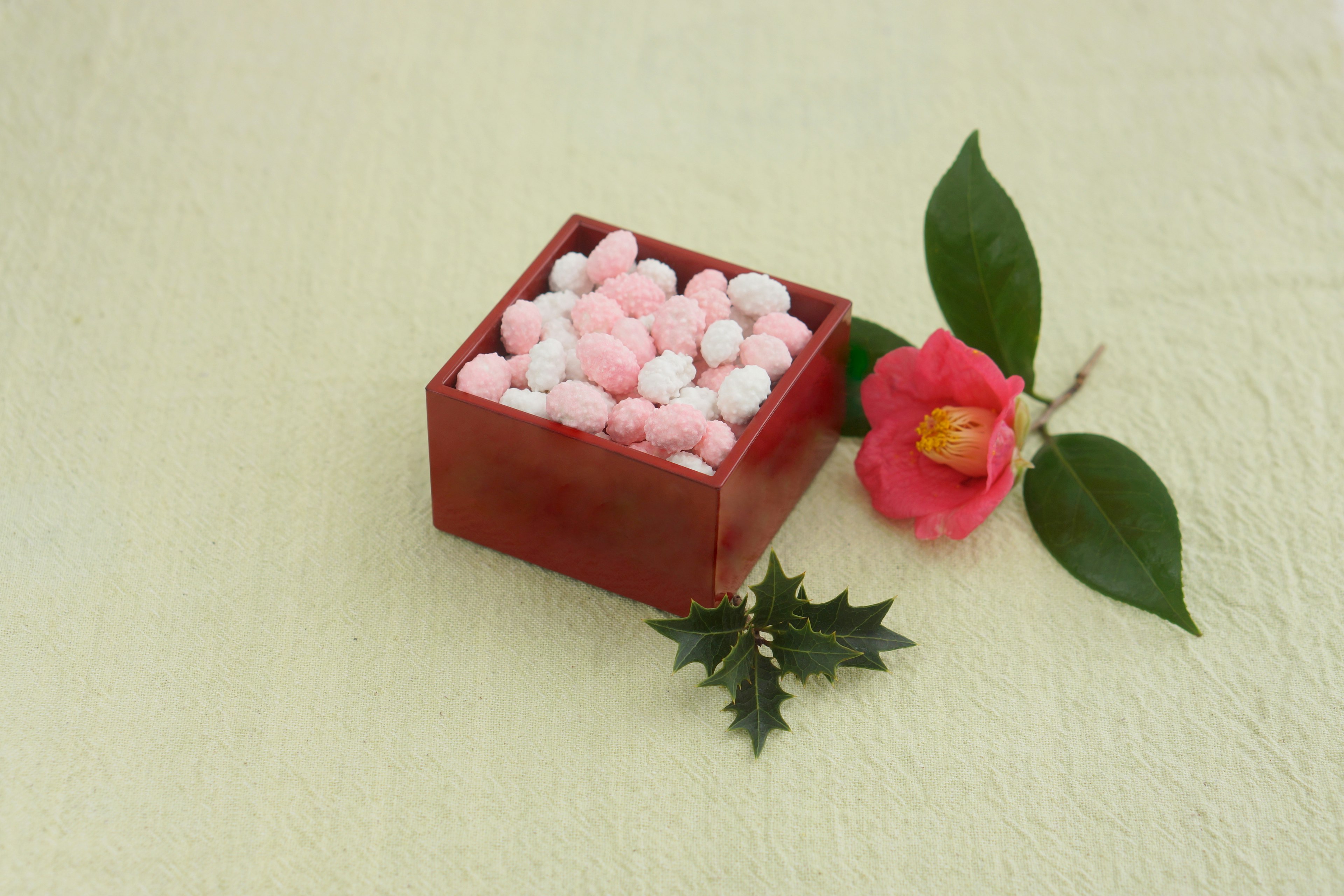 Una caja roja llena de caramelos redondos rosas y blancos junto a una flor de camelia y hojas dispuestas ordenadamente