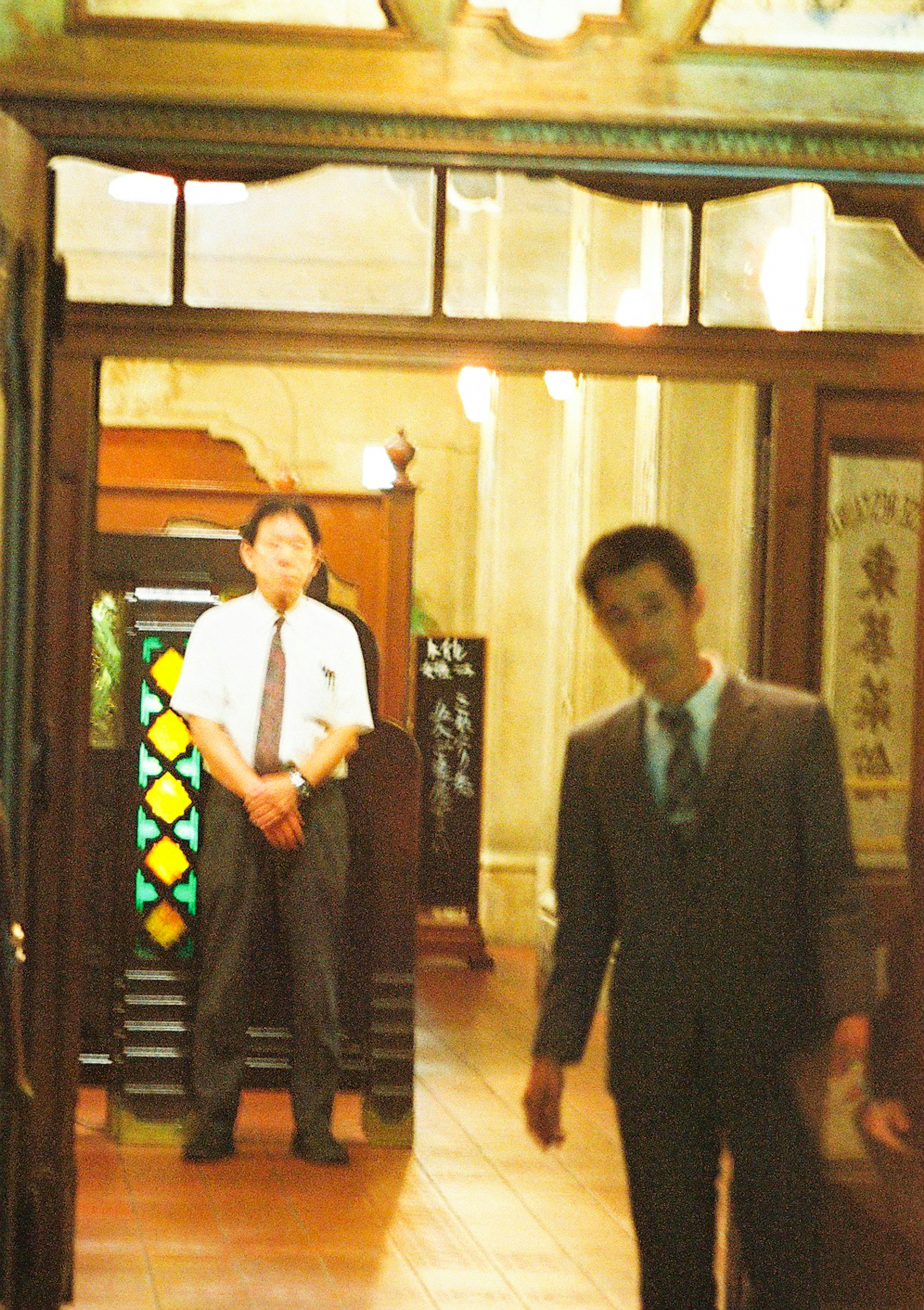 Interior photo featuring two men One man in a suit is walking while another man in a white shirt is standing Colorful stained glass is visible in the background