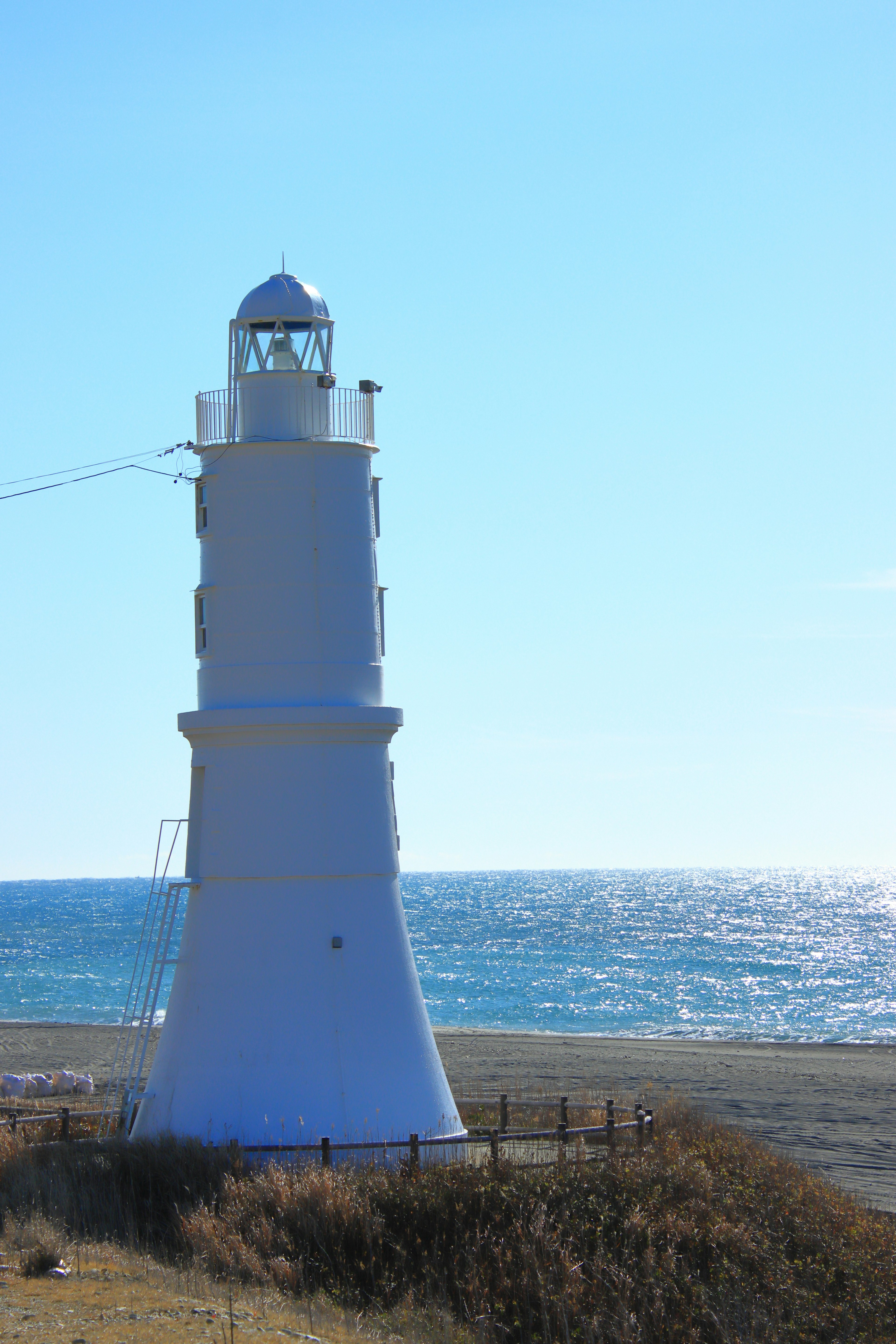 Ein weißer Leuchtturm steht nahe dem Meer unter einem blauen Himmel und ruhigen Gewässern