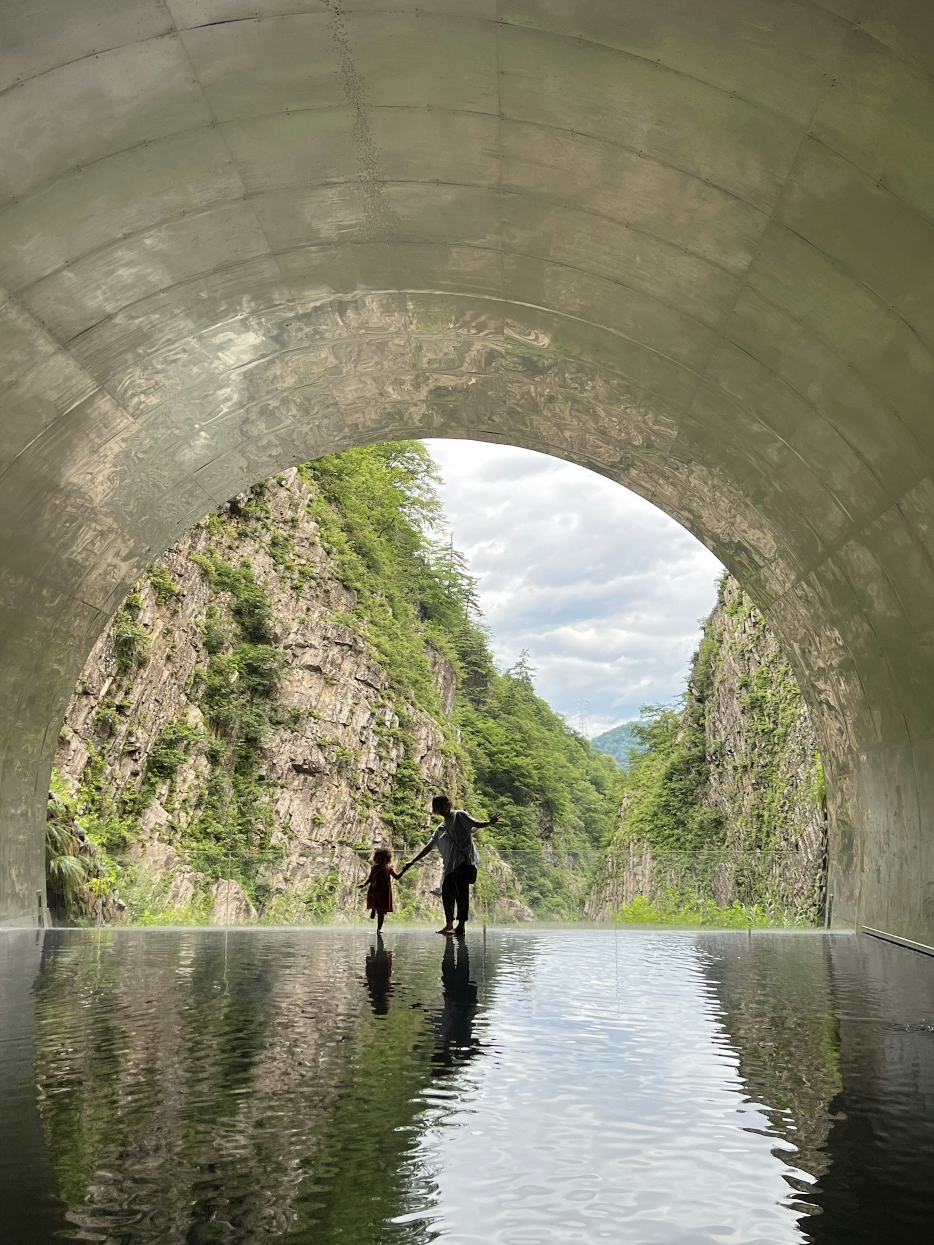 A parent and child standing inside a tunnel Reflection on the water Green mountains in the background