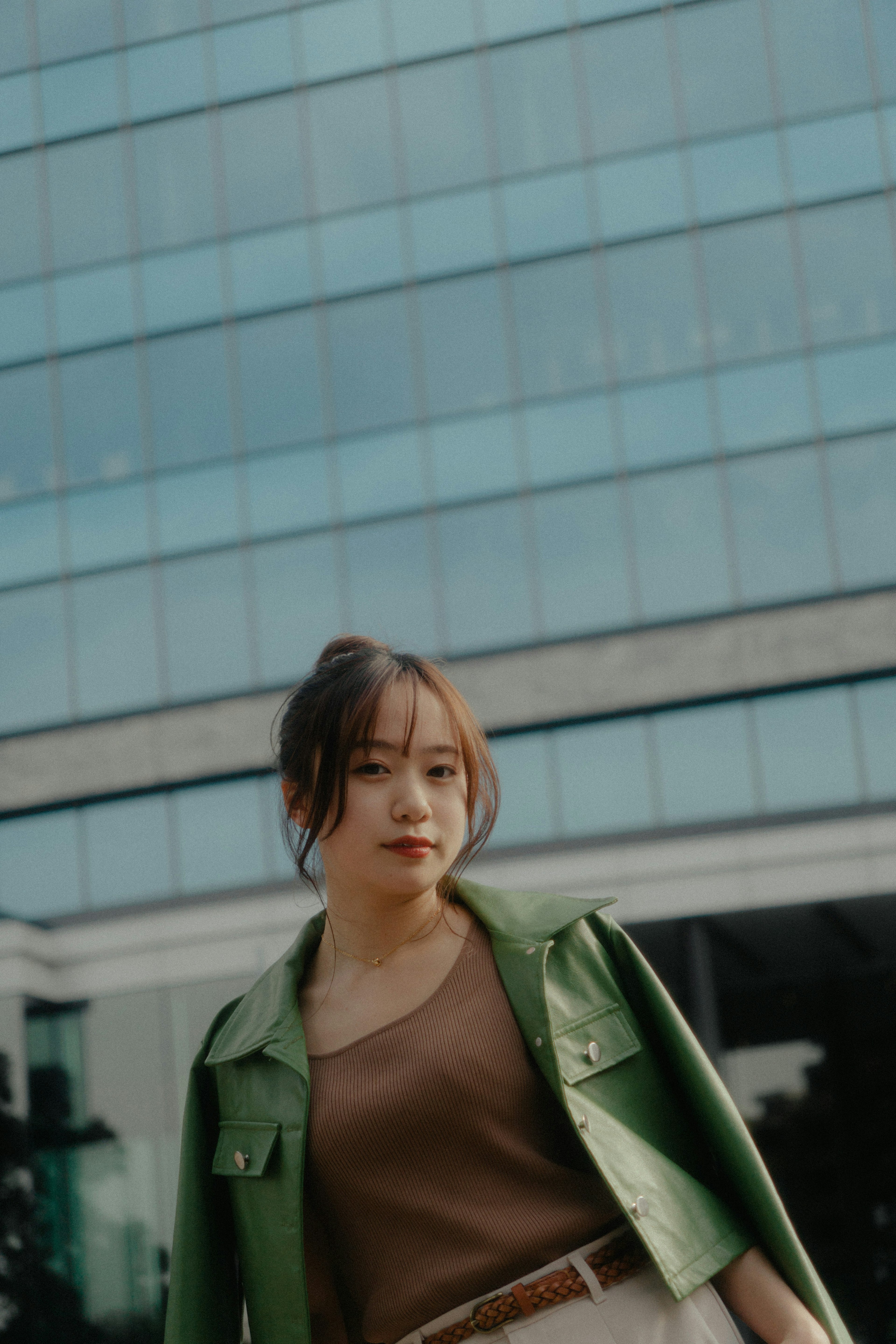 A woman wearing a green jacket with a brown top standing in front of a glass building