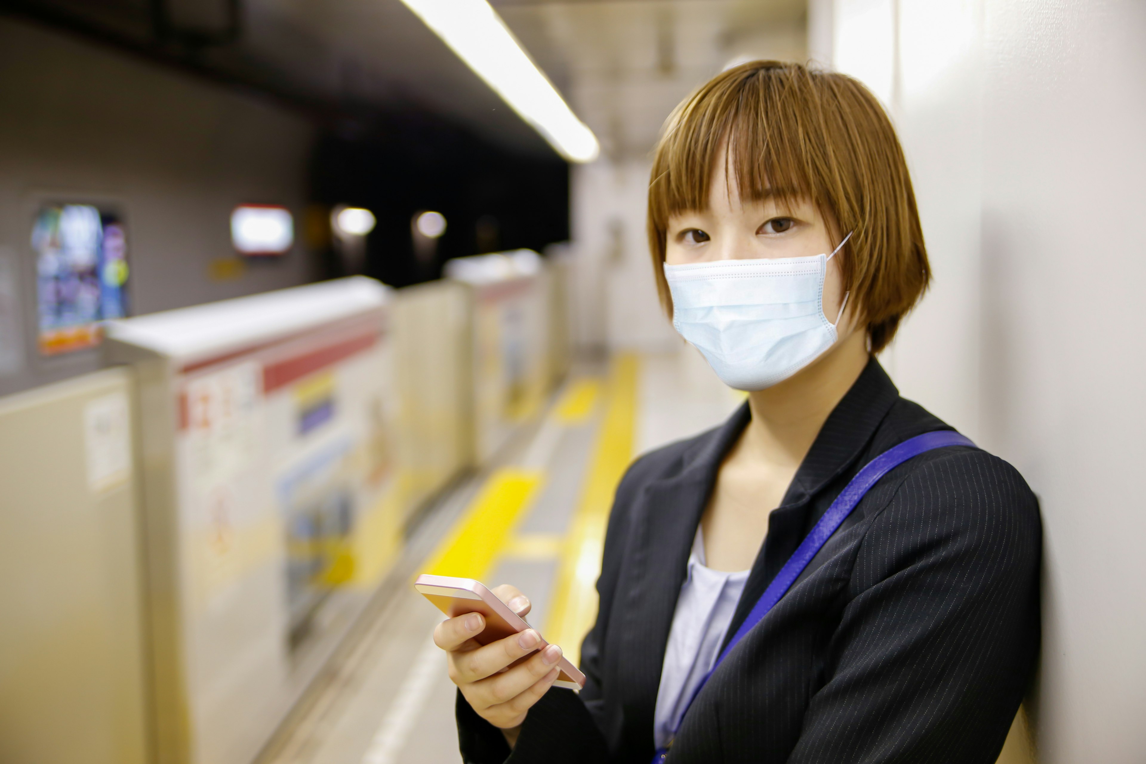 マスクを着用した女性がスマートフォンを持ち、駅のプラットフォームに立っている