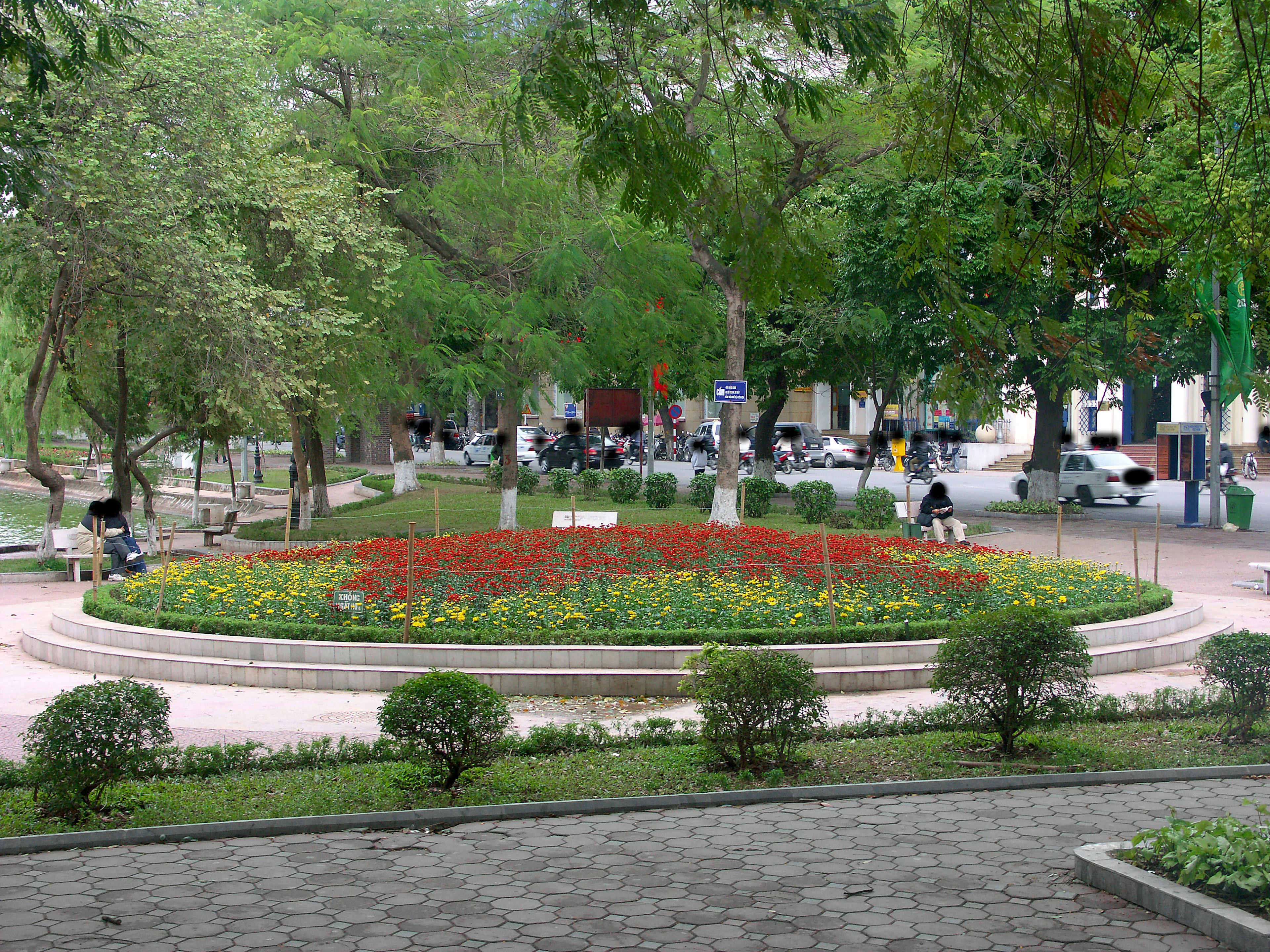 Aiuola circolare in un parco con fiori colorati circondata da alberi verdi e panchine