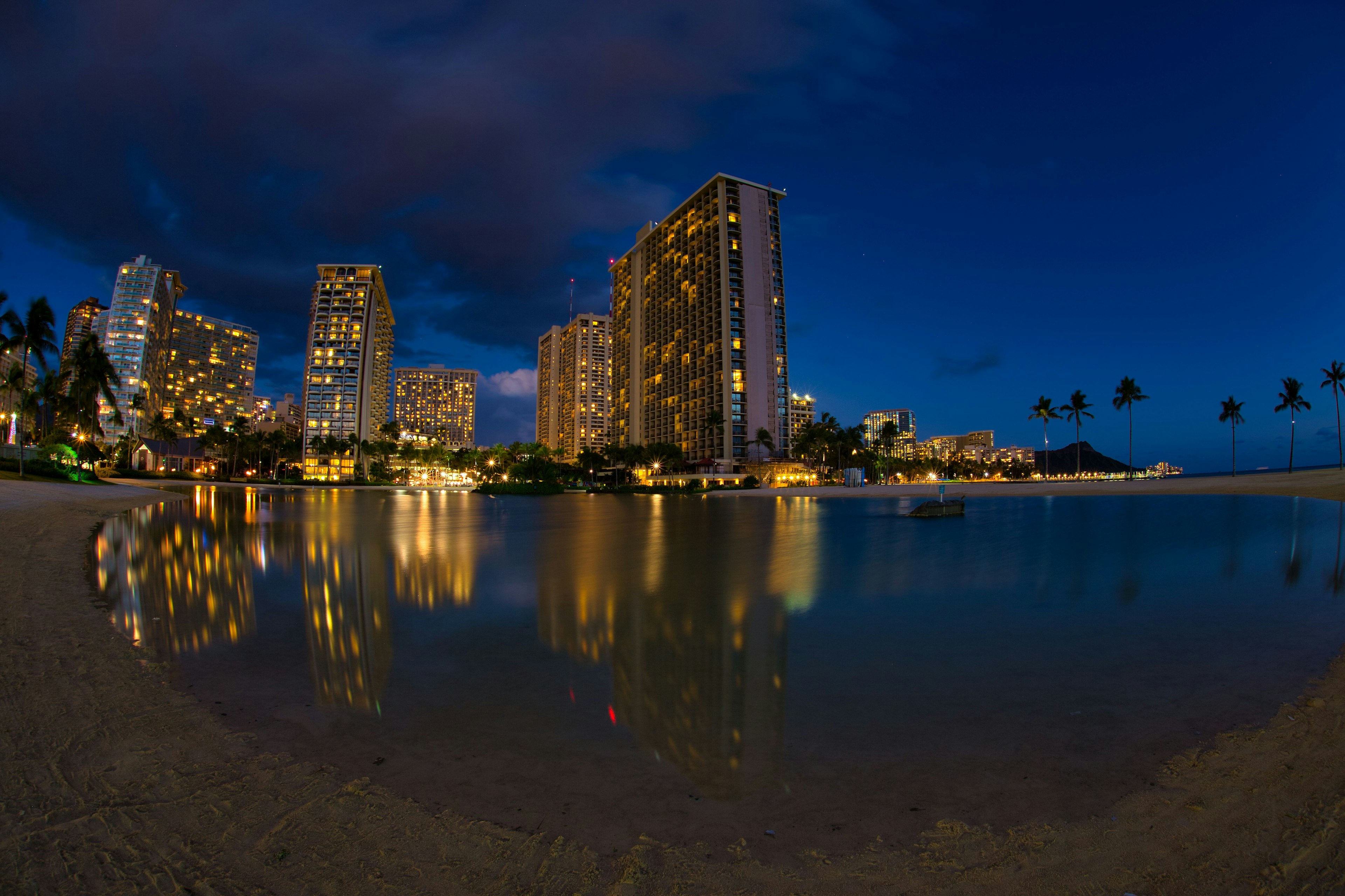 Pantai Waikiki di malam hari dengan gedung pencakar langit dan refleksi di air