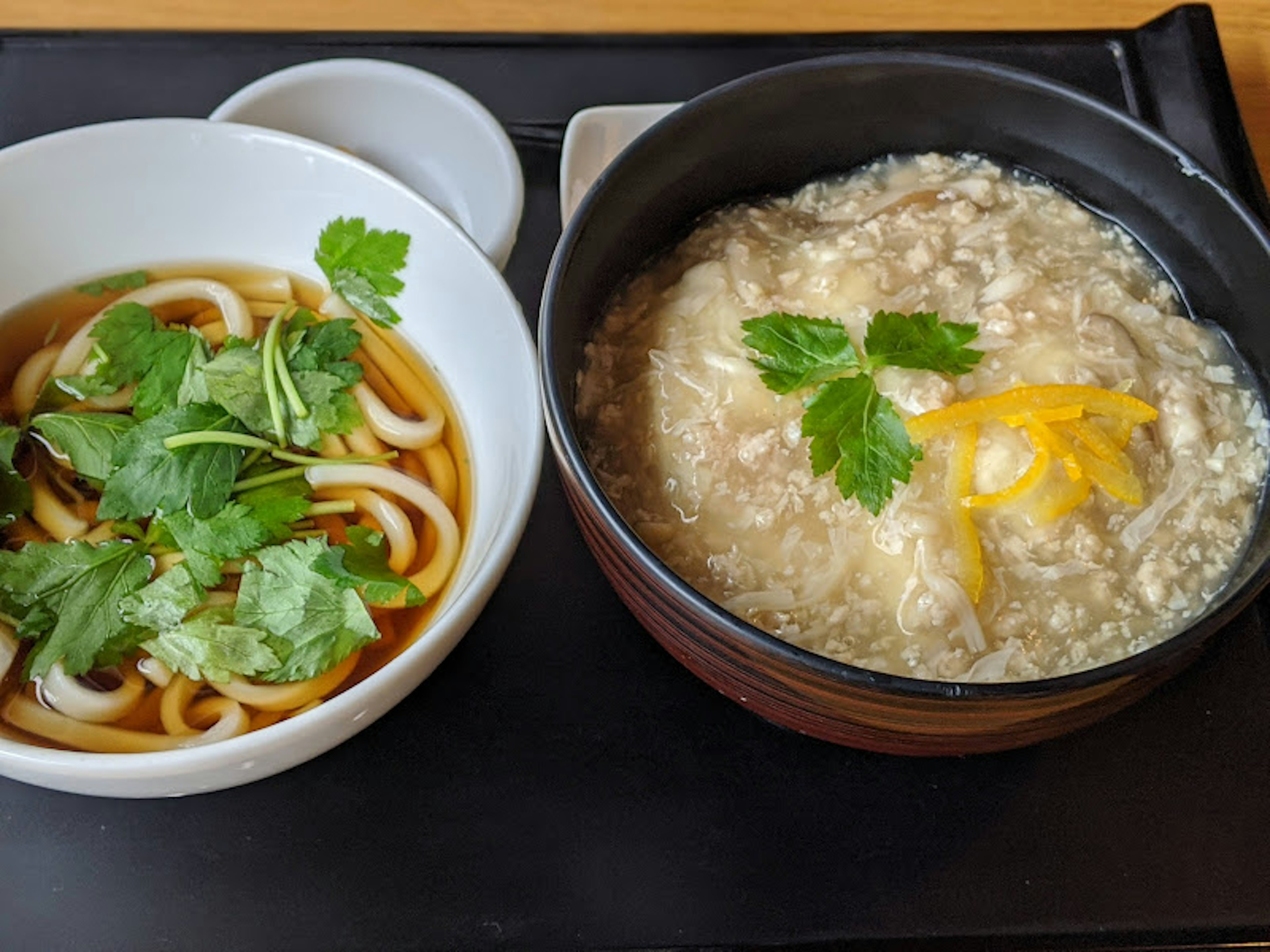 白いボウルに入ったうどんと、黒いボウルの粥に香菜とレモンがトッピングされた料理