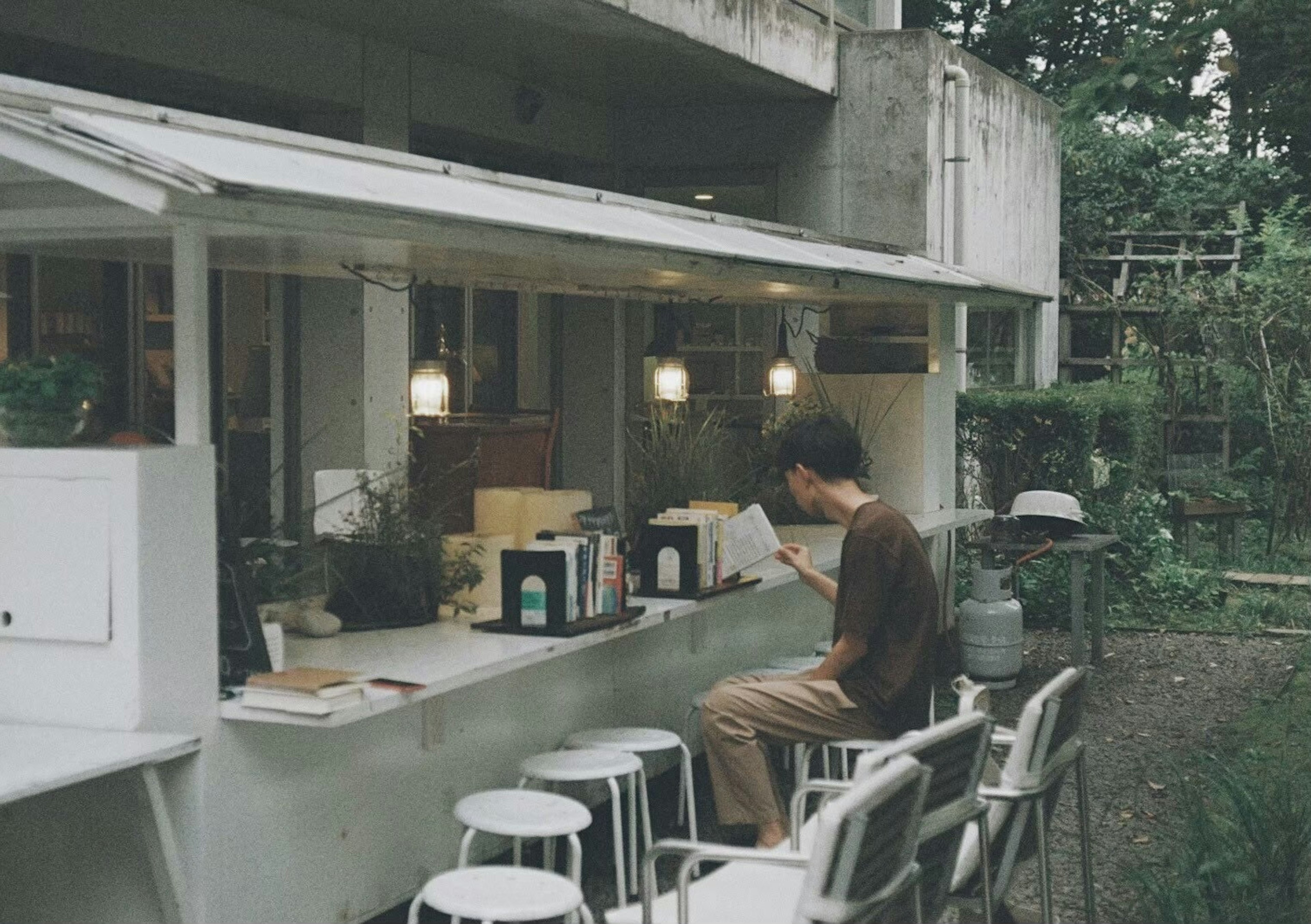 Mann liest ein Buch auf der Terrasse eines Cafés umgeben von Pflanzen und einfacher Dekoration