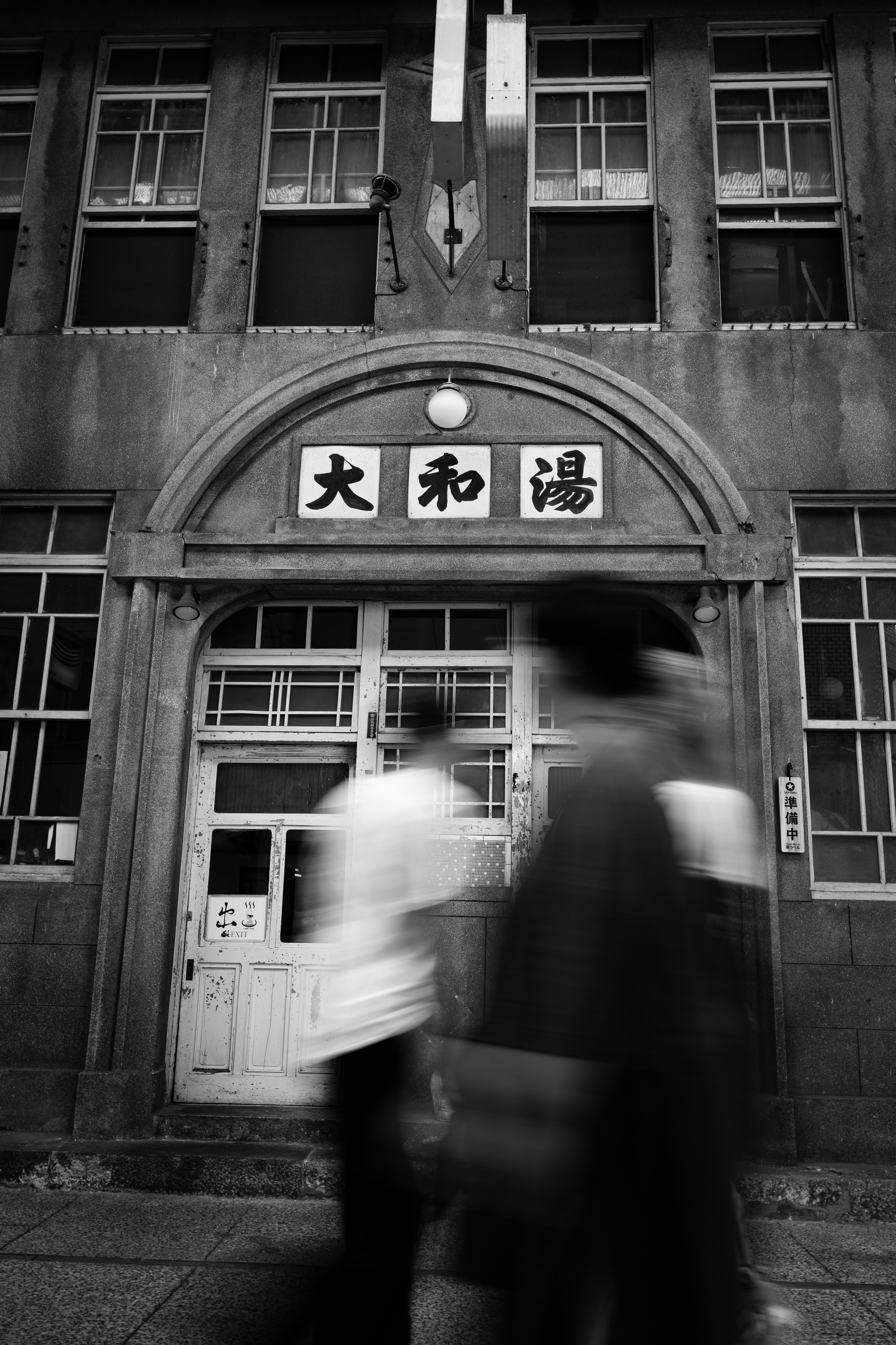 Personas caminando frente a un edificio antiguo en blanco y negro con un letrero que dice Daiwa-yu