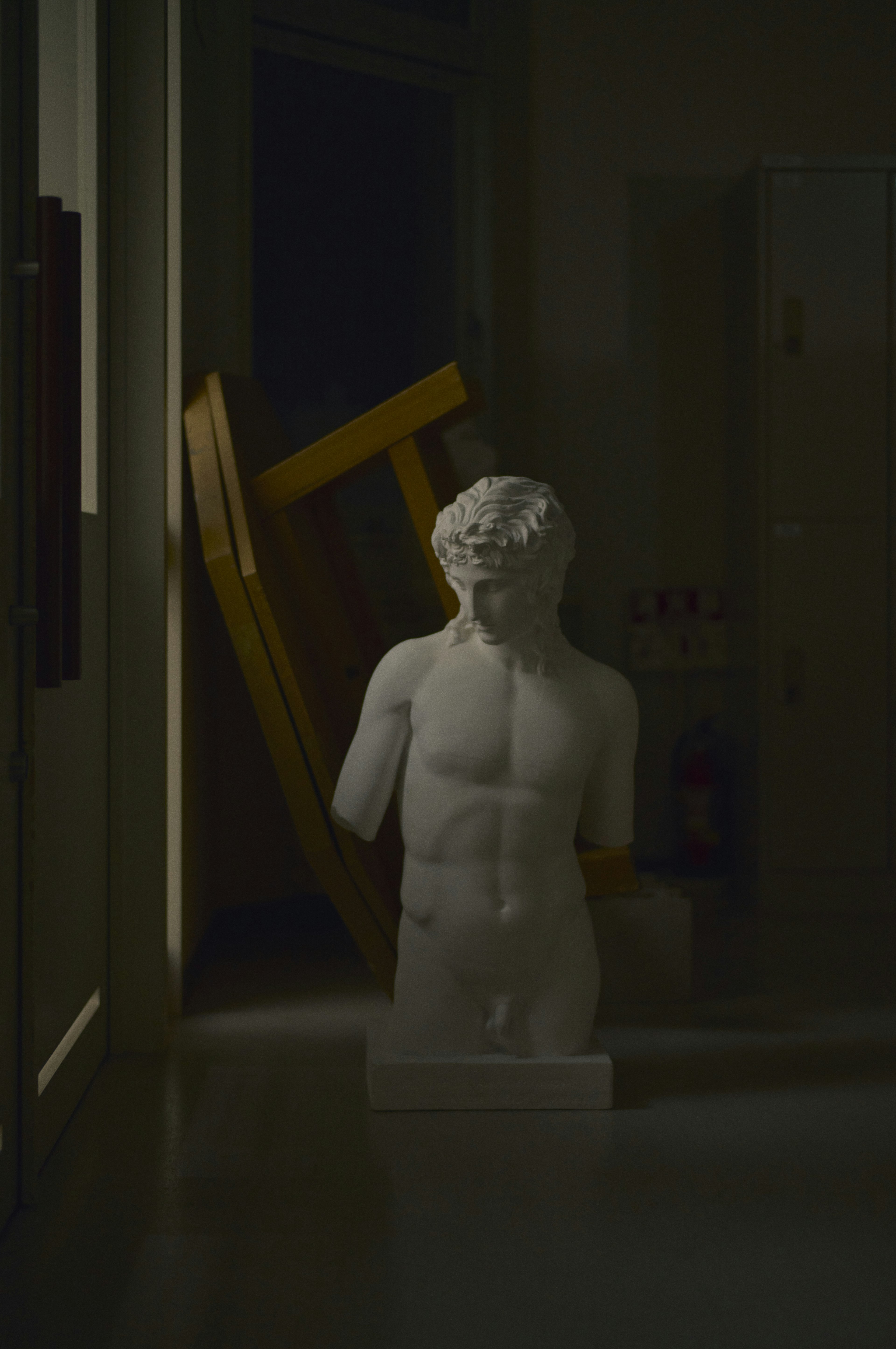 White male sculpture in a dimly lit room with a fallen yellow chair in the background