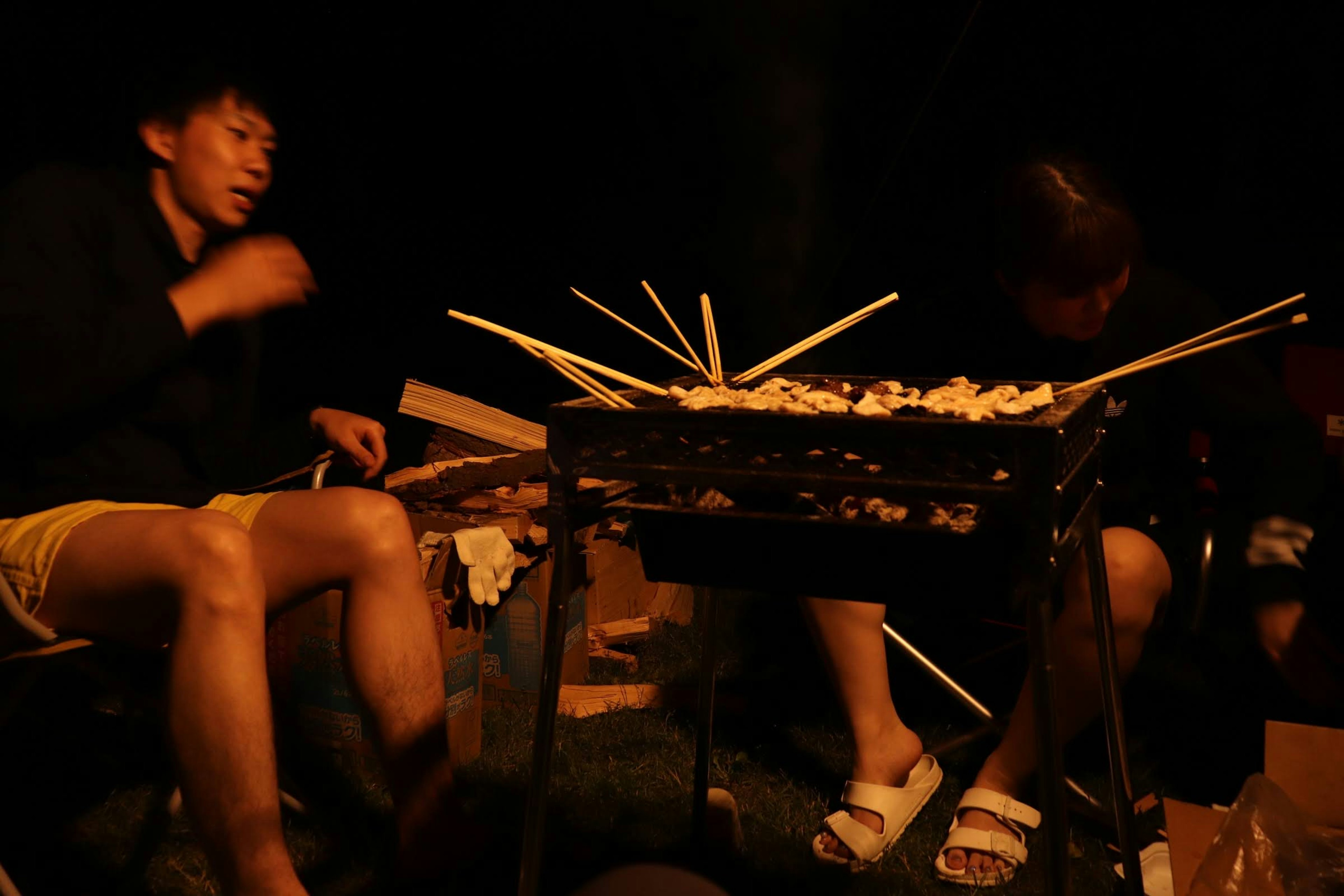 Dos personas asando comida en brochetas en un ambiente oscuro