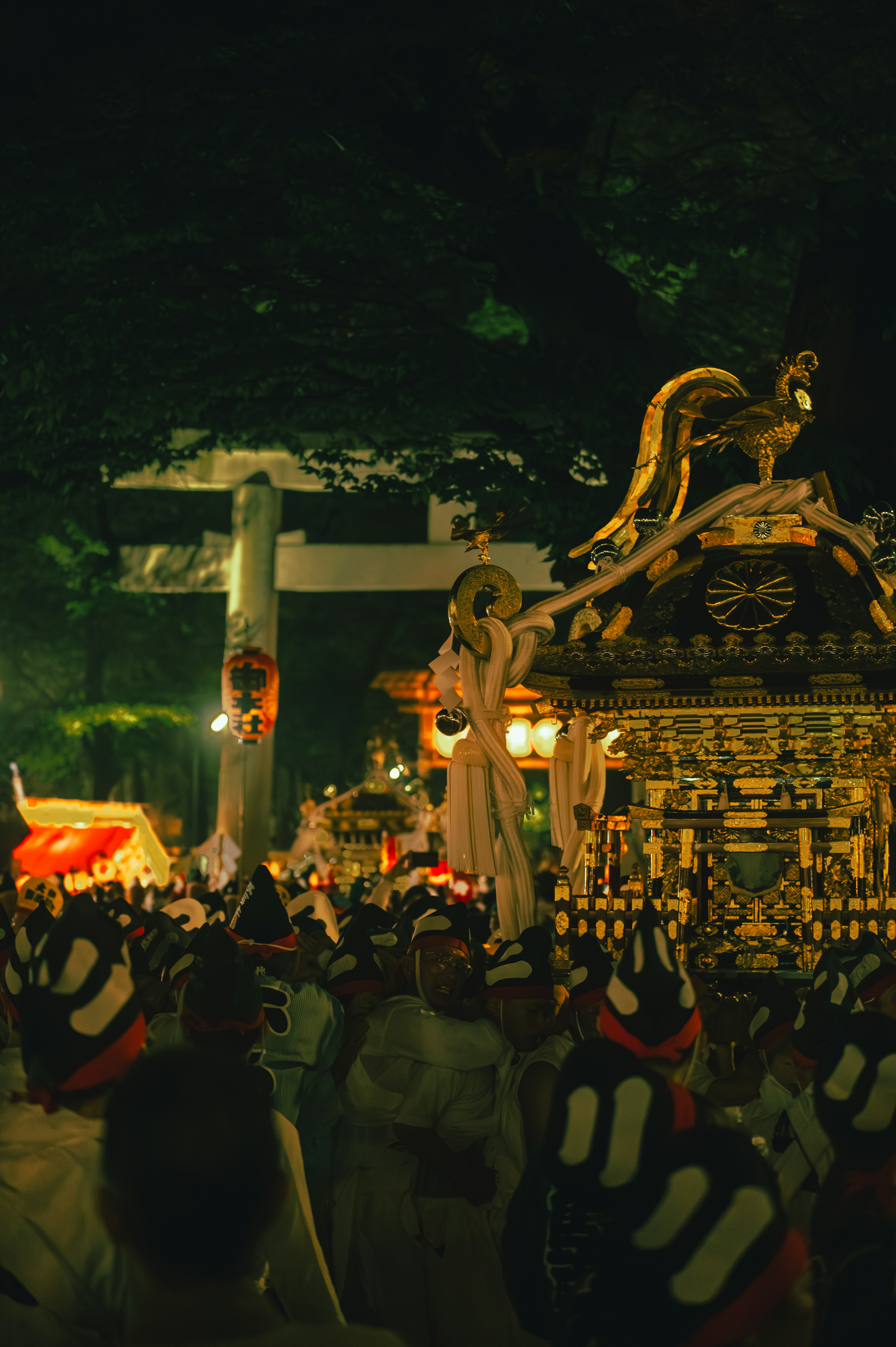 Cảnh lễ hội ban đêm với mikoshi được bao quanh bởi mọi người và cổng torii ở phía sau