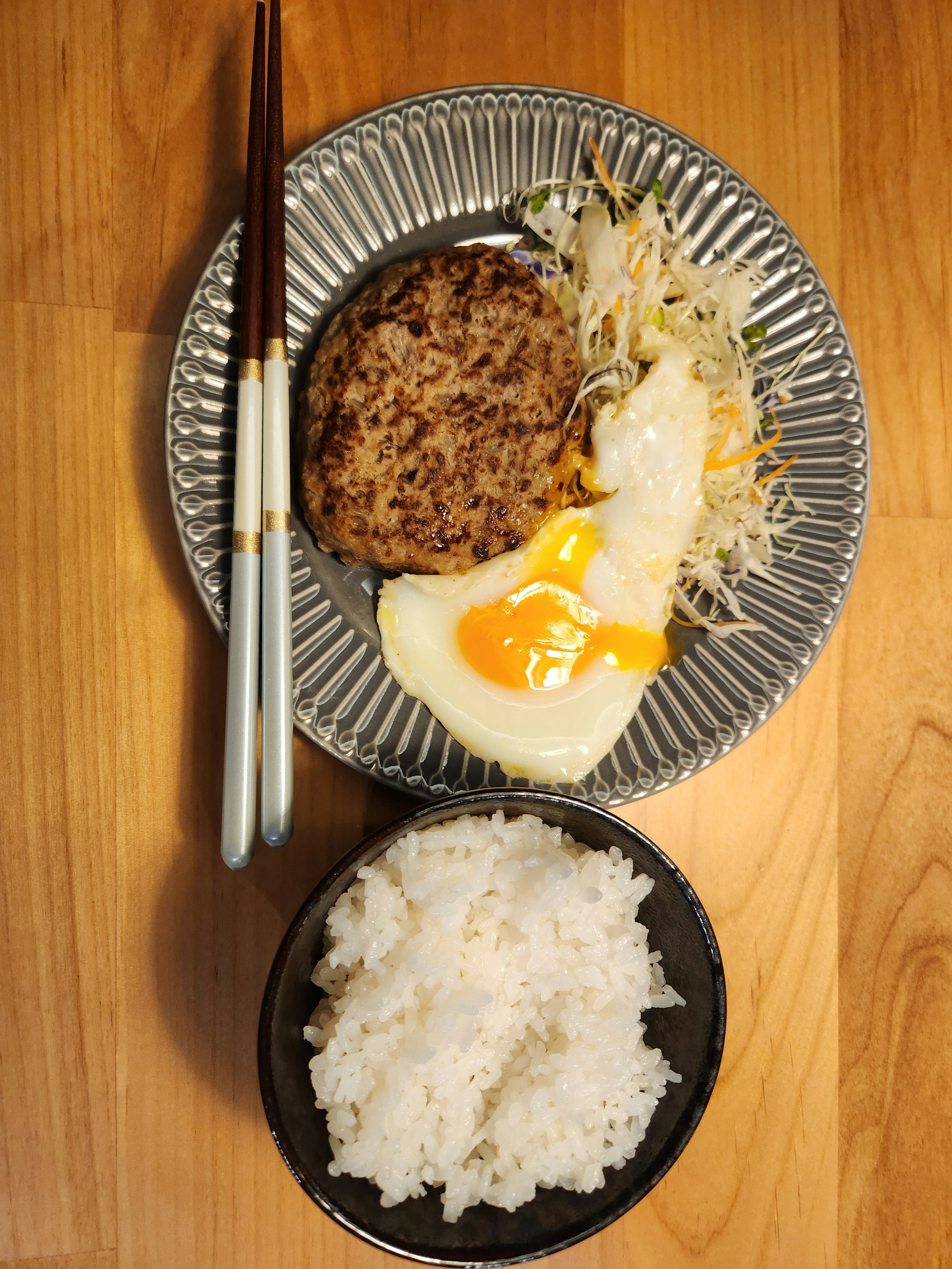 A plate with hamburger steak, fried egg, and cabbage salad alongside a bowl of rice