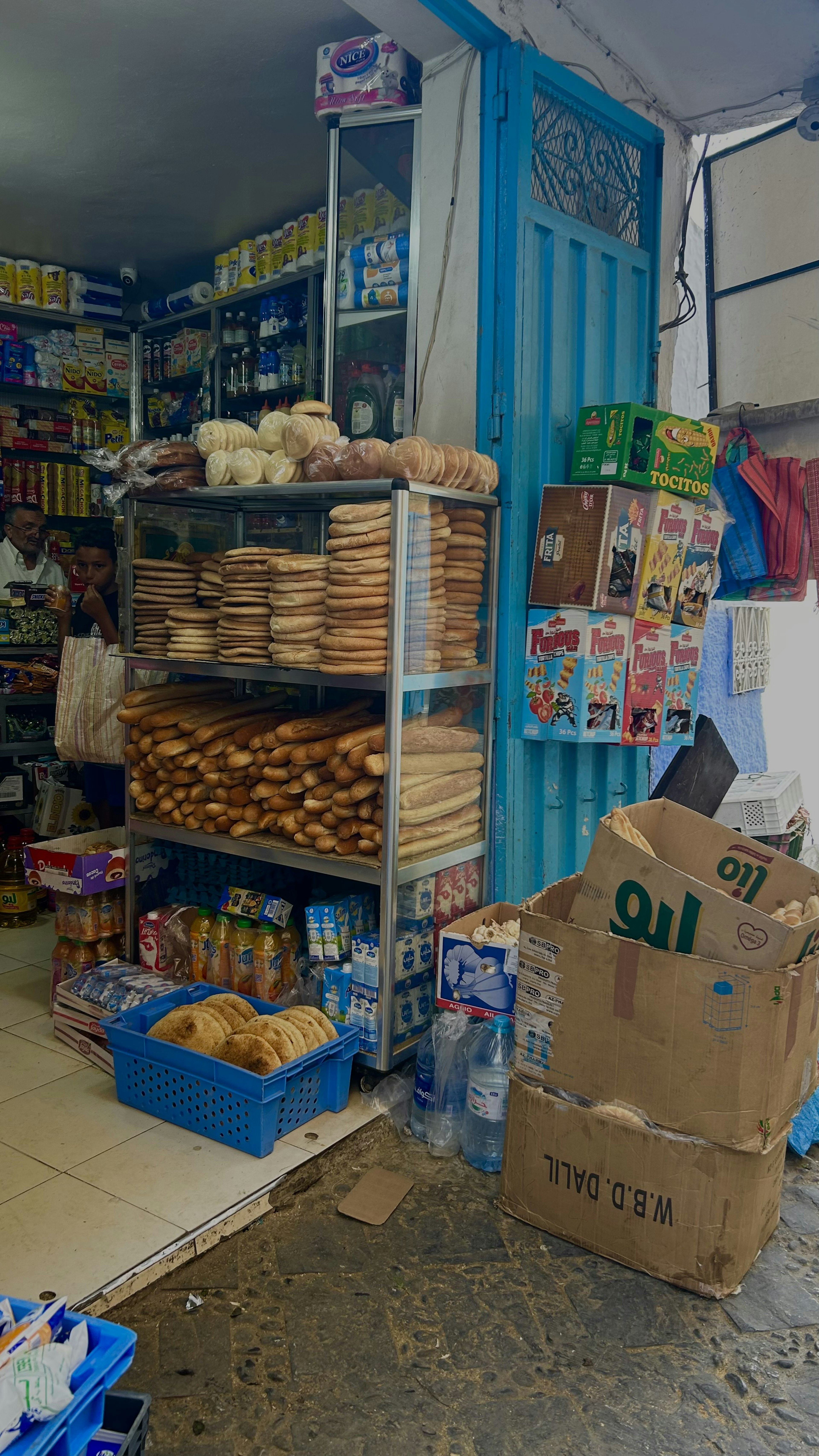 Interior de una pequeña tienda con estantes de pan y comestibles contra una pared azul