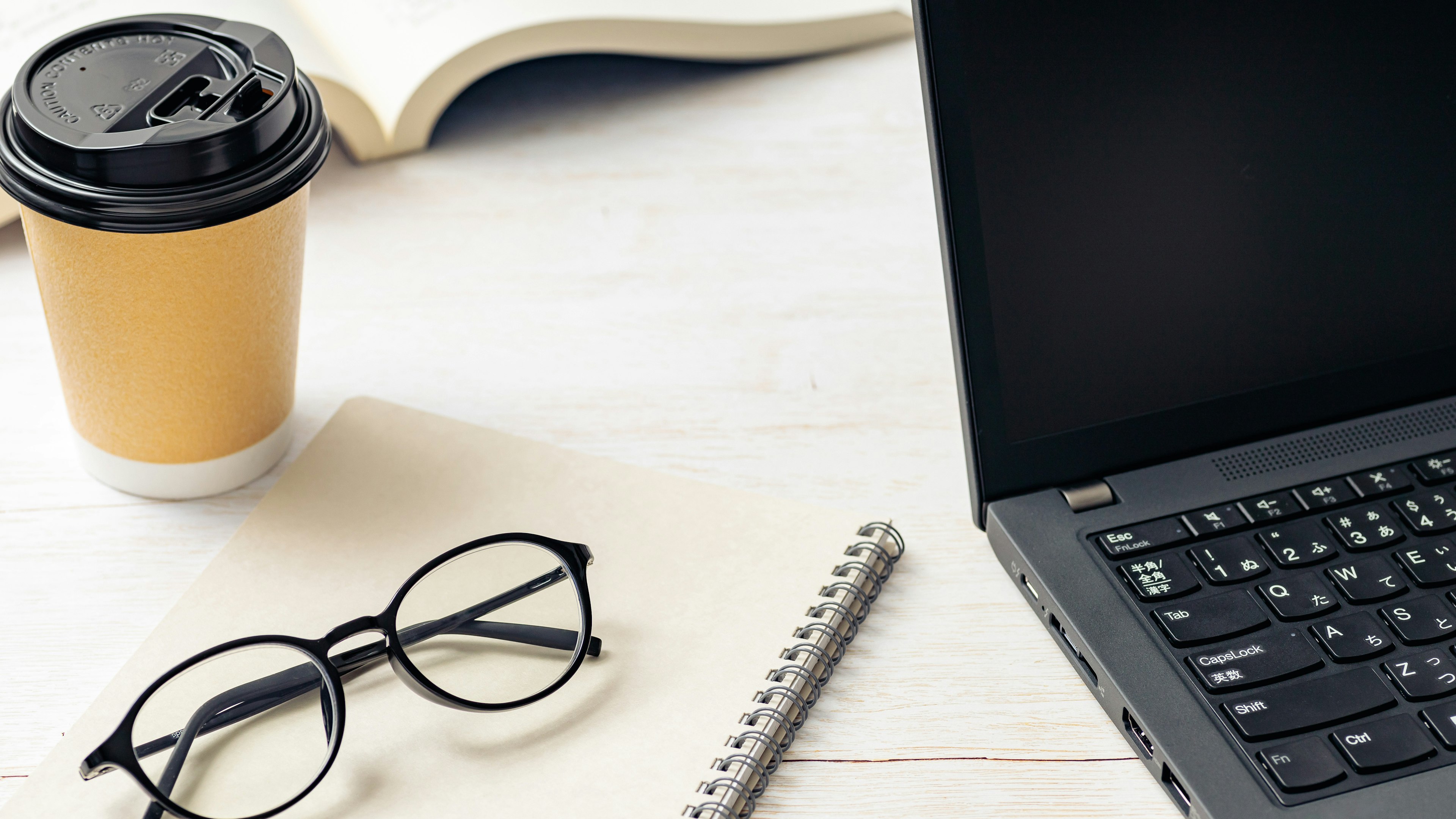 A laptop and coffee cup on a desk with glasses and a notebook nearby