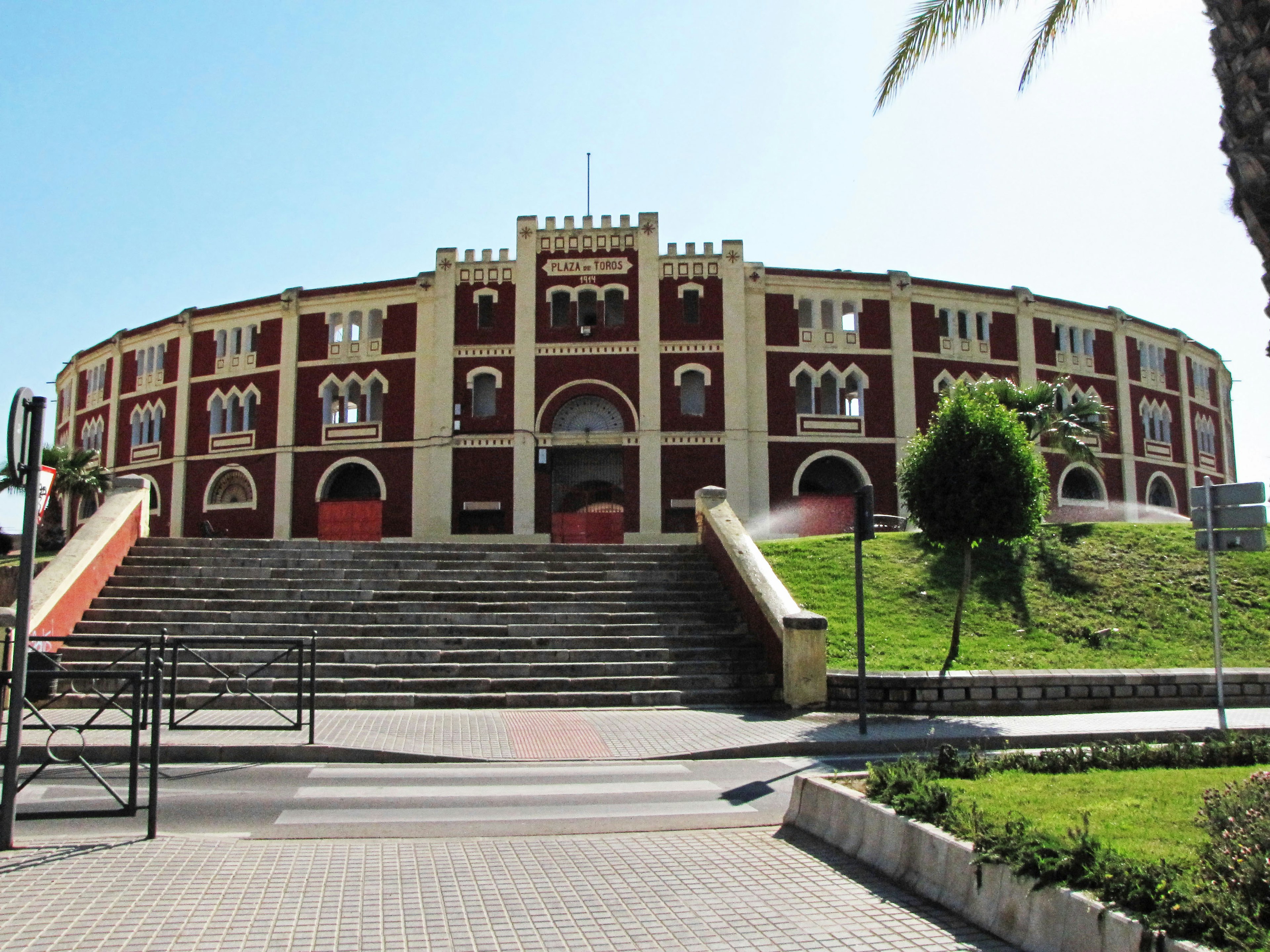 Vista exterior de una arena, edificio rojo y blanco, escaleras, paisajismo verde