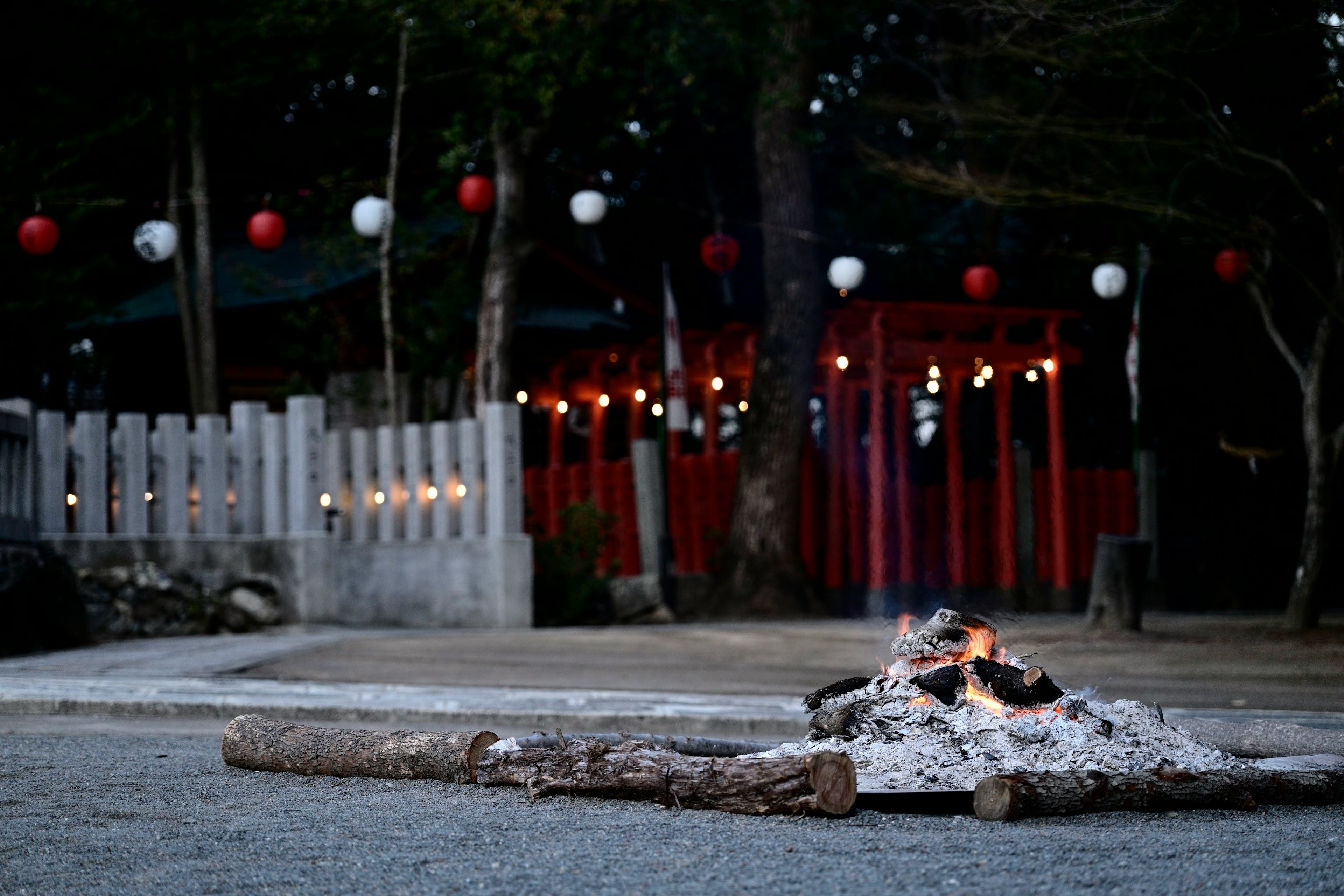 地面上燃燒的木頭和灰燼，模糊的燈籠和神社背景