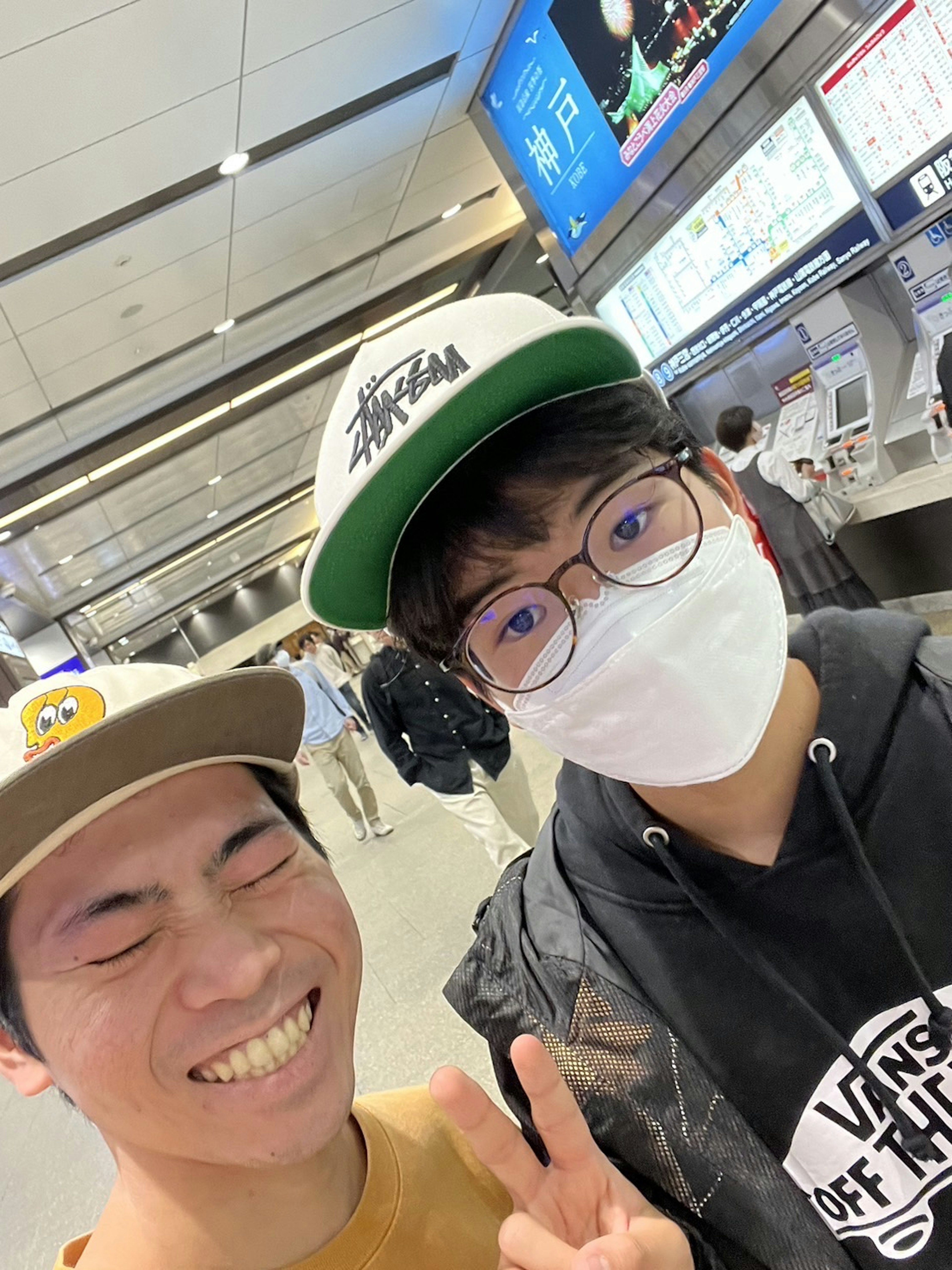 Two young men taking a selfie in a train station one smiling and giving a peace sign while the other wears a mask