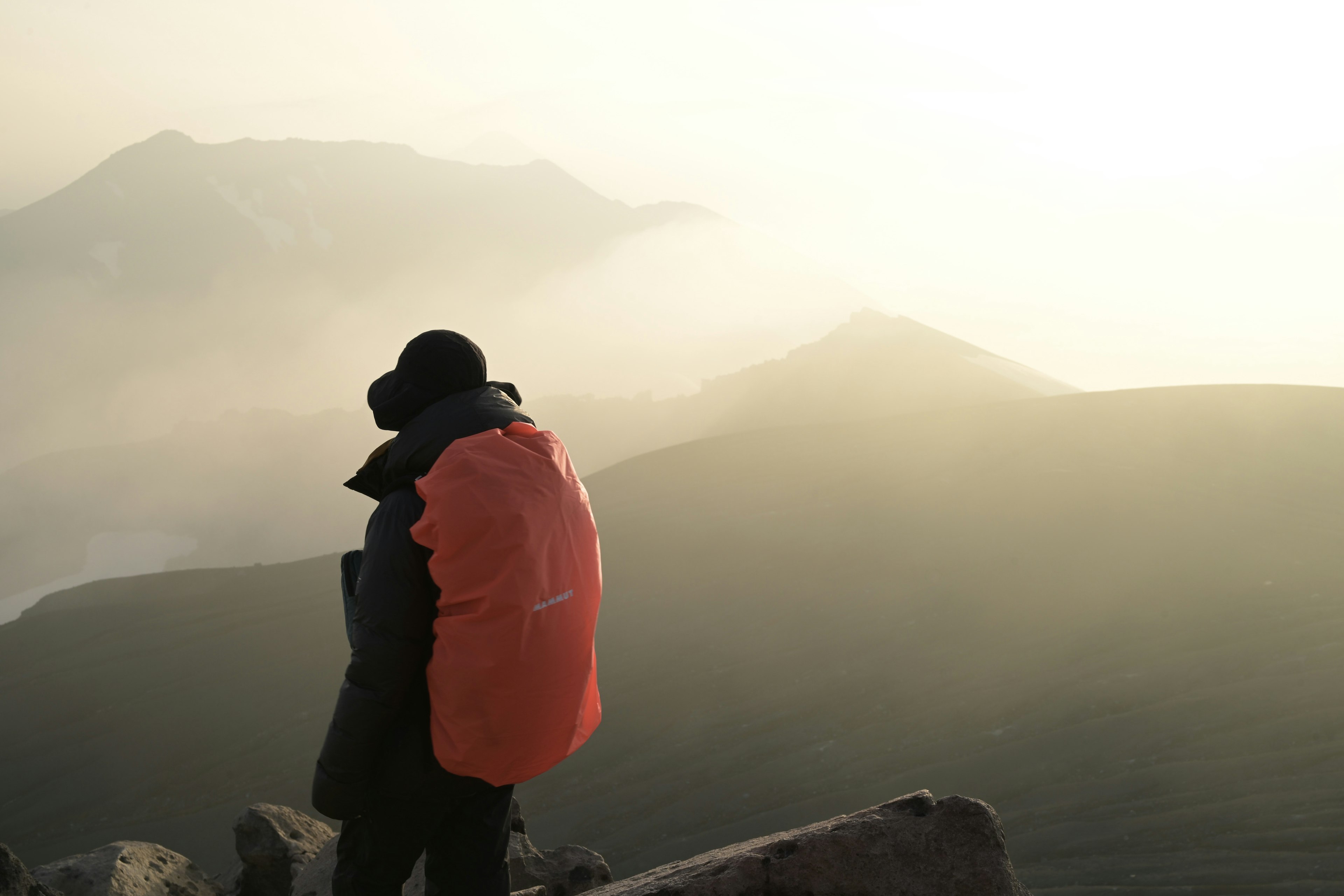 Eine Person steht am Berggipfel mit einem orangefarbenen Rucksack und nebligen Bergen im Hintergrund