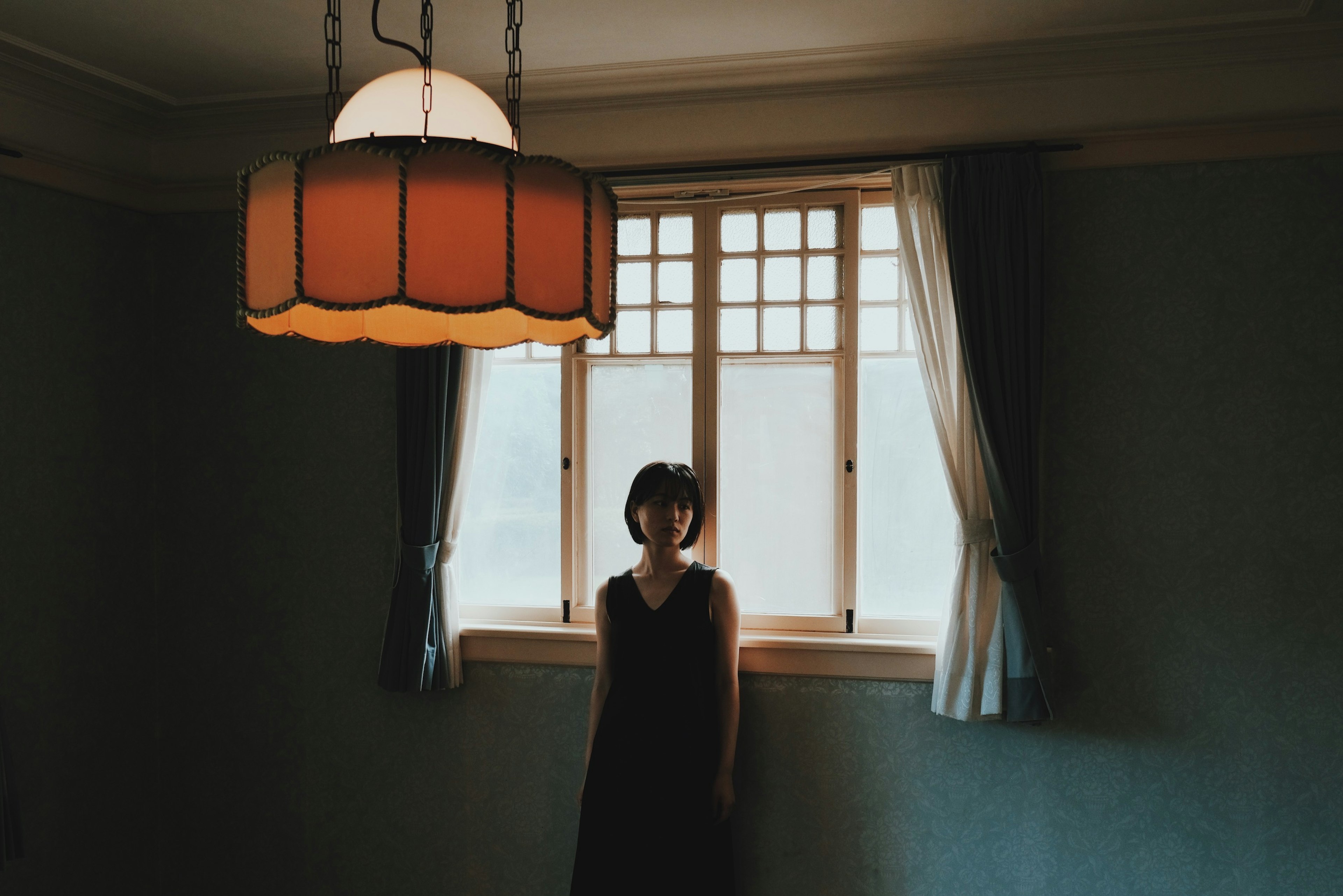 A woman stands in a dim room with an orange lamp hanging above