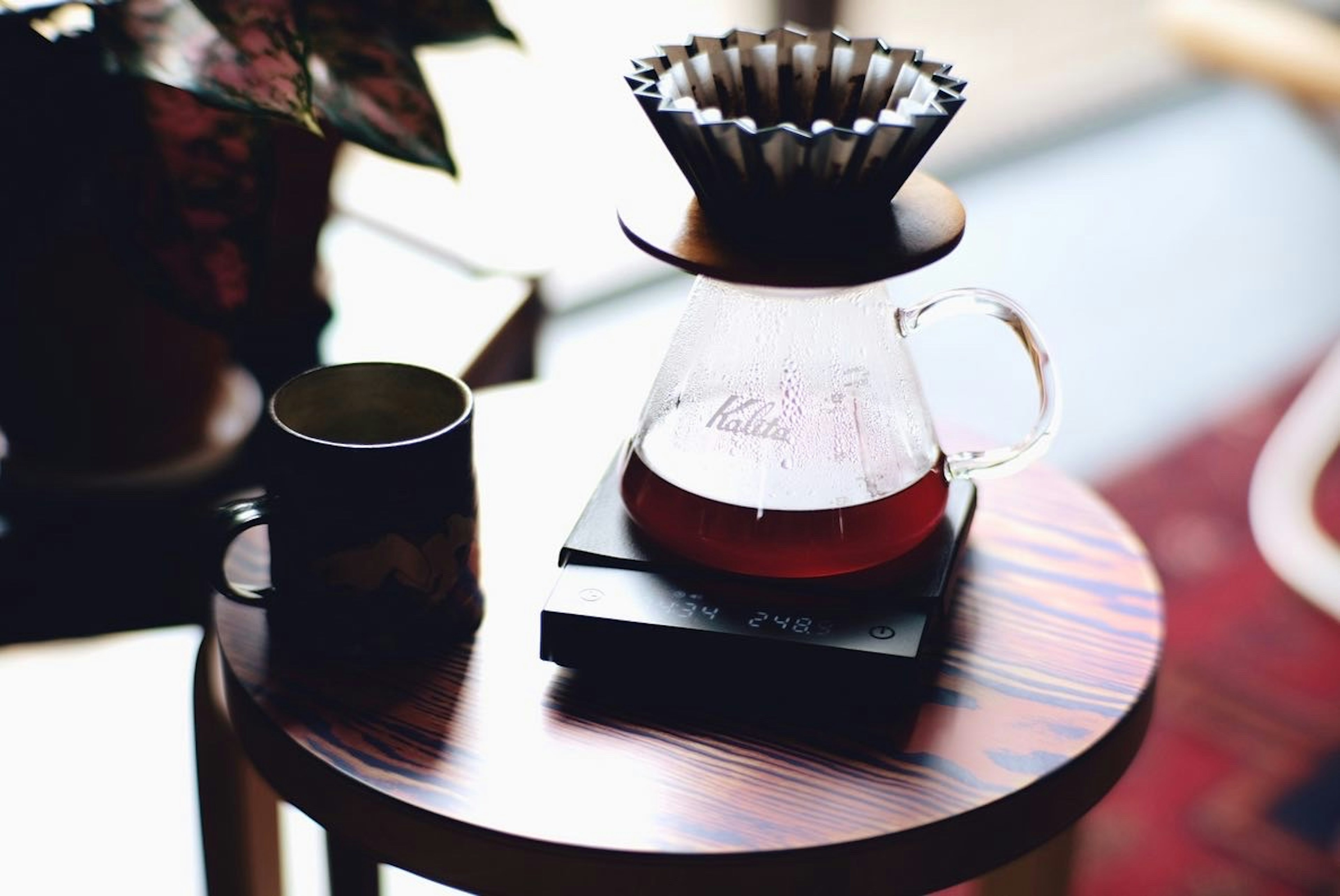 A coffee dripper and glass server on a wooden table with a cup and plant