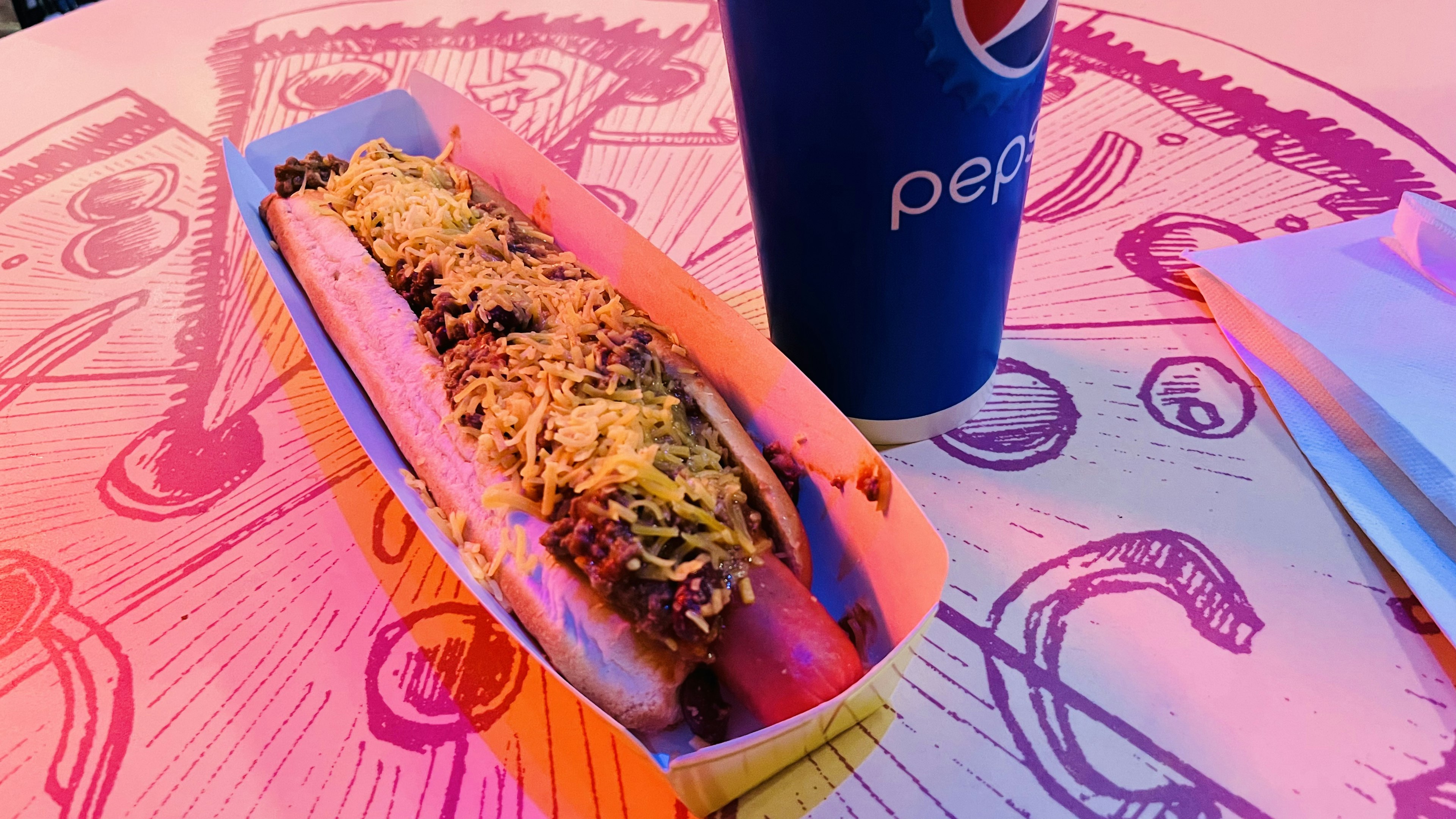 Un hot dog garni de fromage à côté d'une tasse de Pepsi sur une table colorée