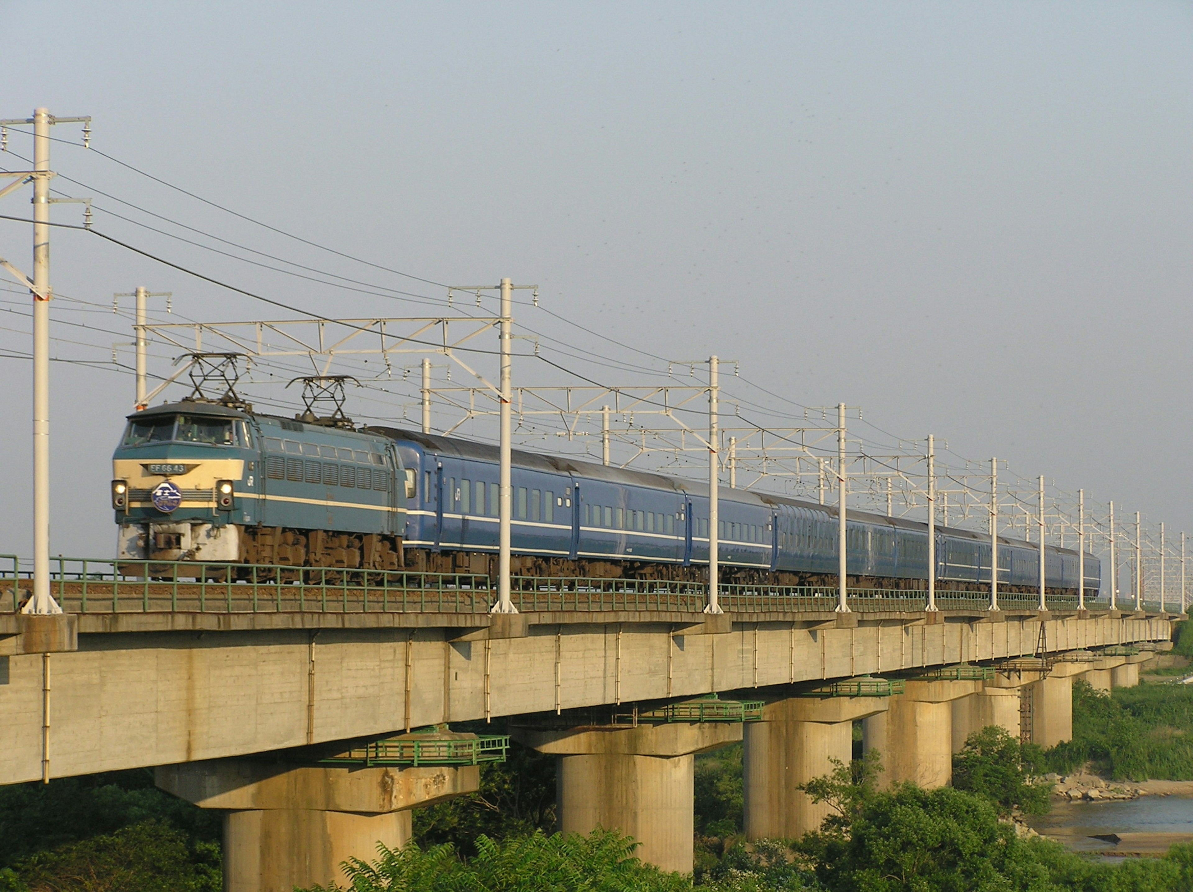 Kereta biru melaju di atas jembatan dengan kabel listrik