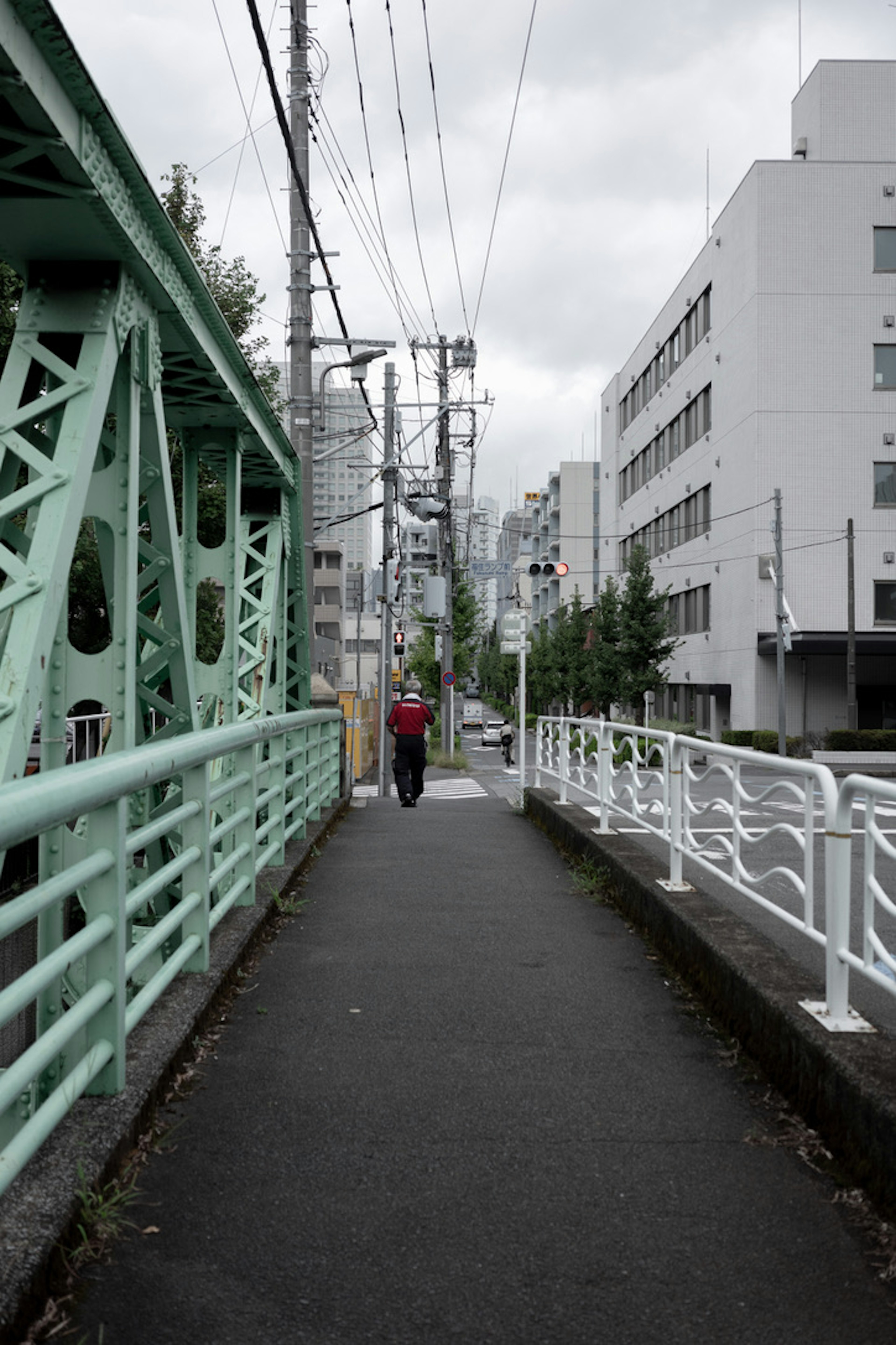 Person walking on a walkway beside a green bridge and surrounding buildings