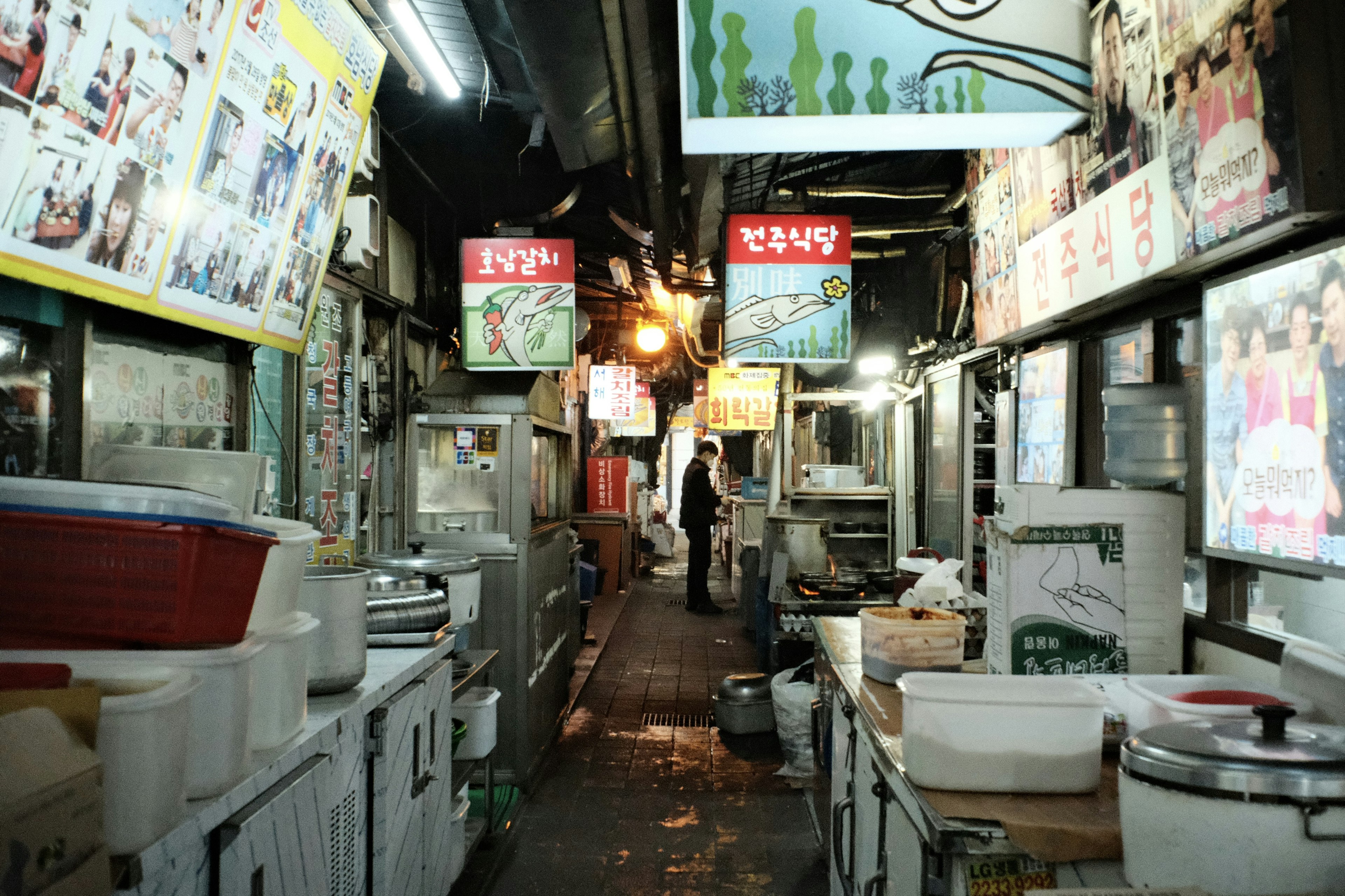 Narrow alley in a street market with shops lined up bright signs visible and a person walking