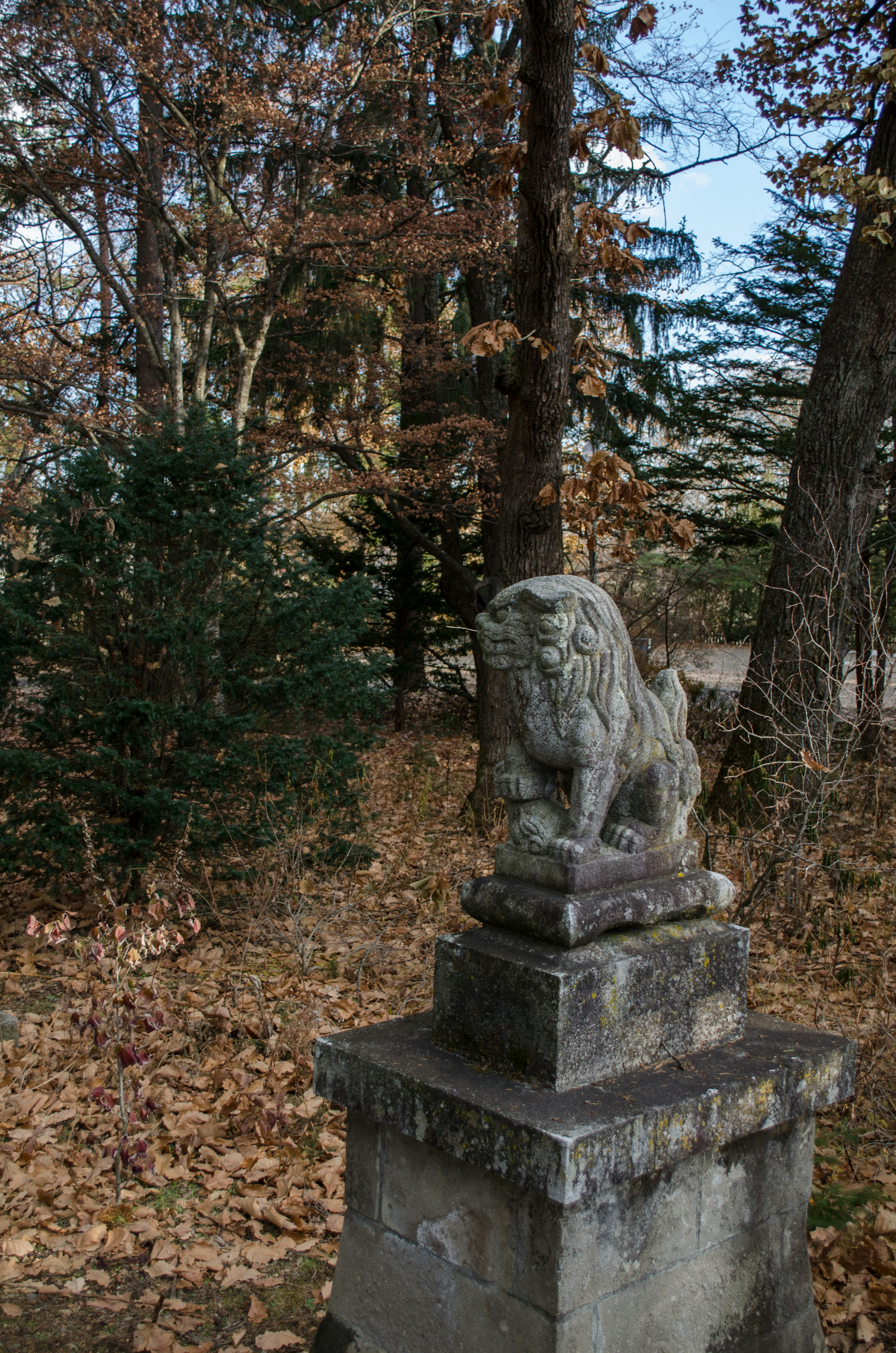 Eine alte Steinstatue eines Löwen steht in einer Landschaft, umgeben von Bäumen und gefallenen Blättern