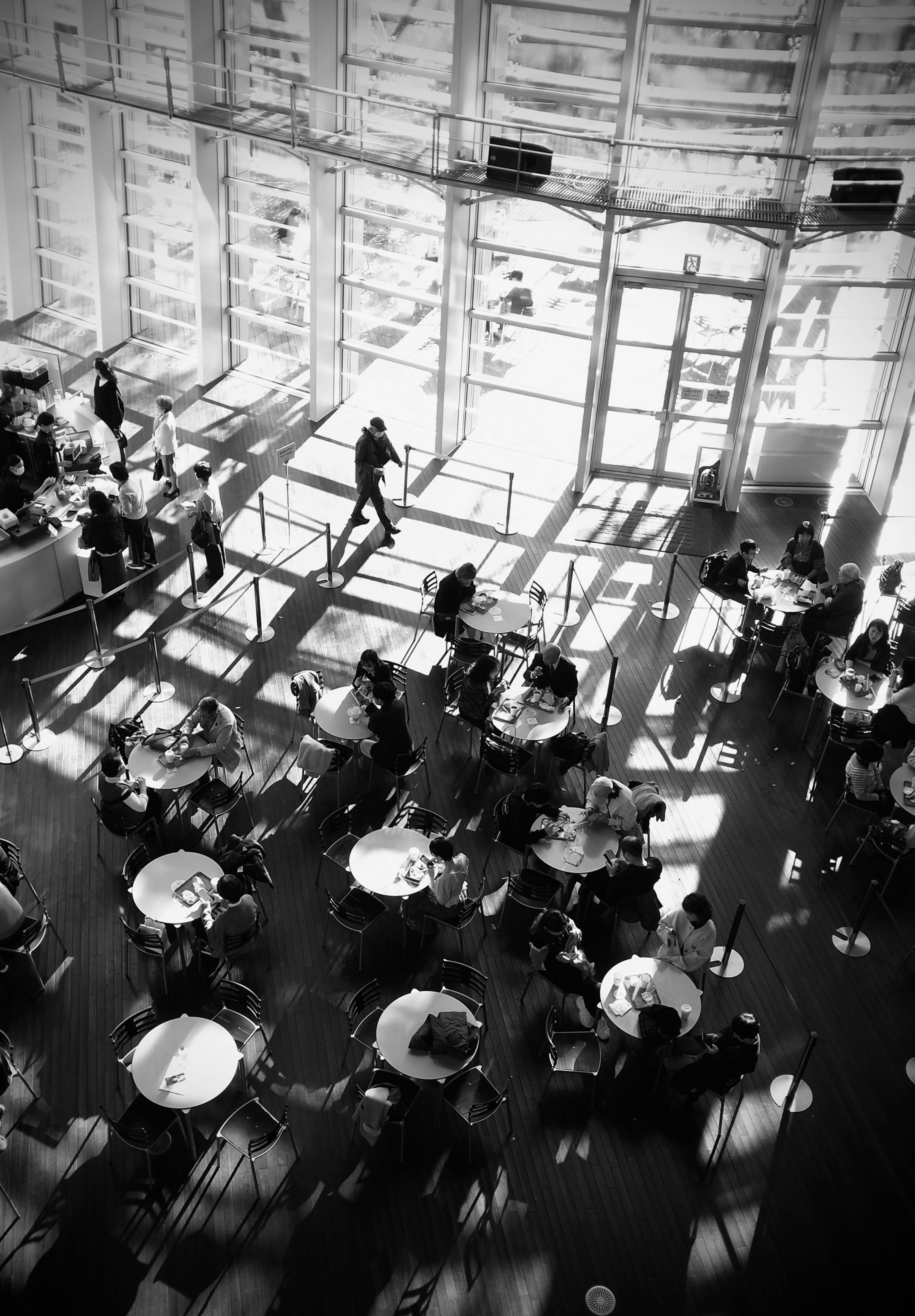 Schwarzweiße Vogelperspektive eines Café-Innenraums mit zahlreichen Tischen und Stühlen und auffälligen Schatten durch natürliches Licht