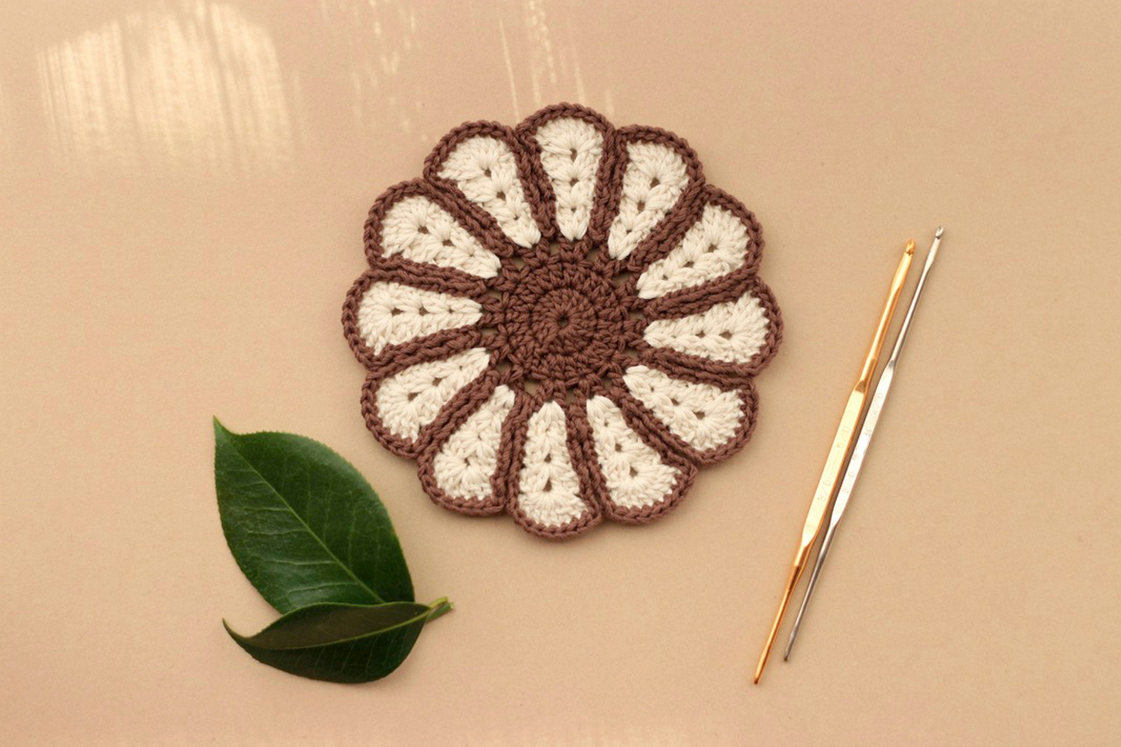 Brown and white flower-shaped crochet coaster with a golden crochet hook and green leaf