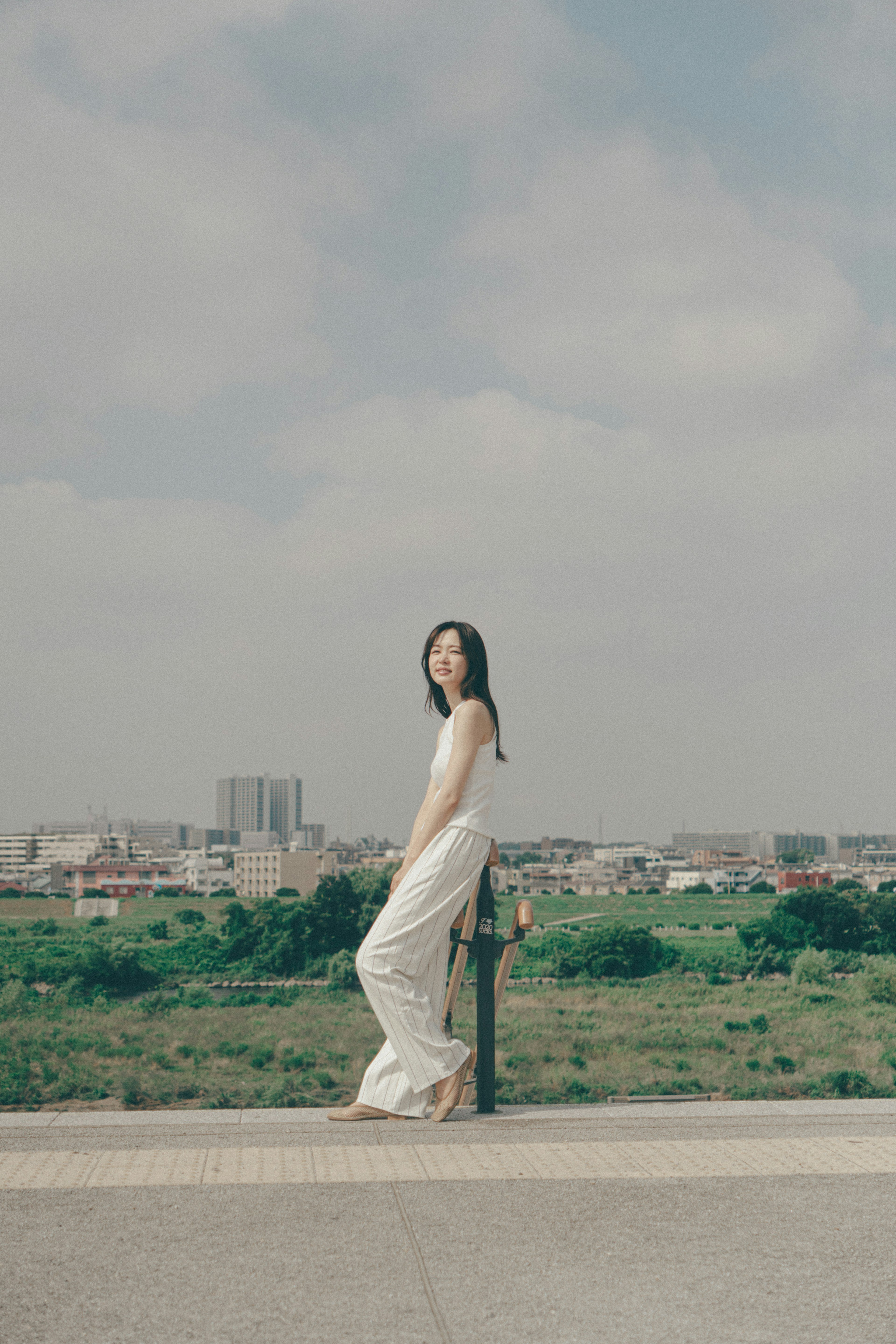 A woman in a white dress stands against a backdrop of urban skyline and green fields