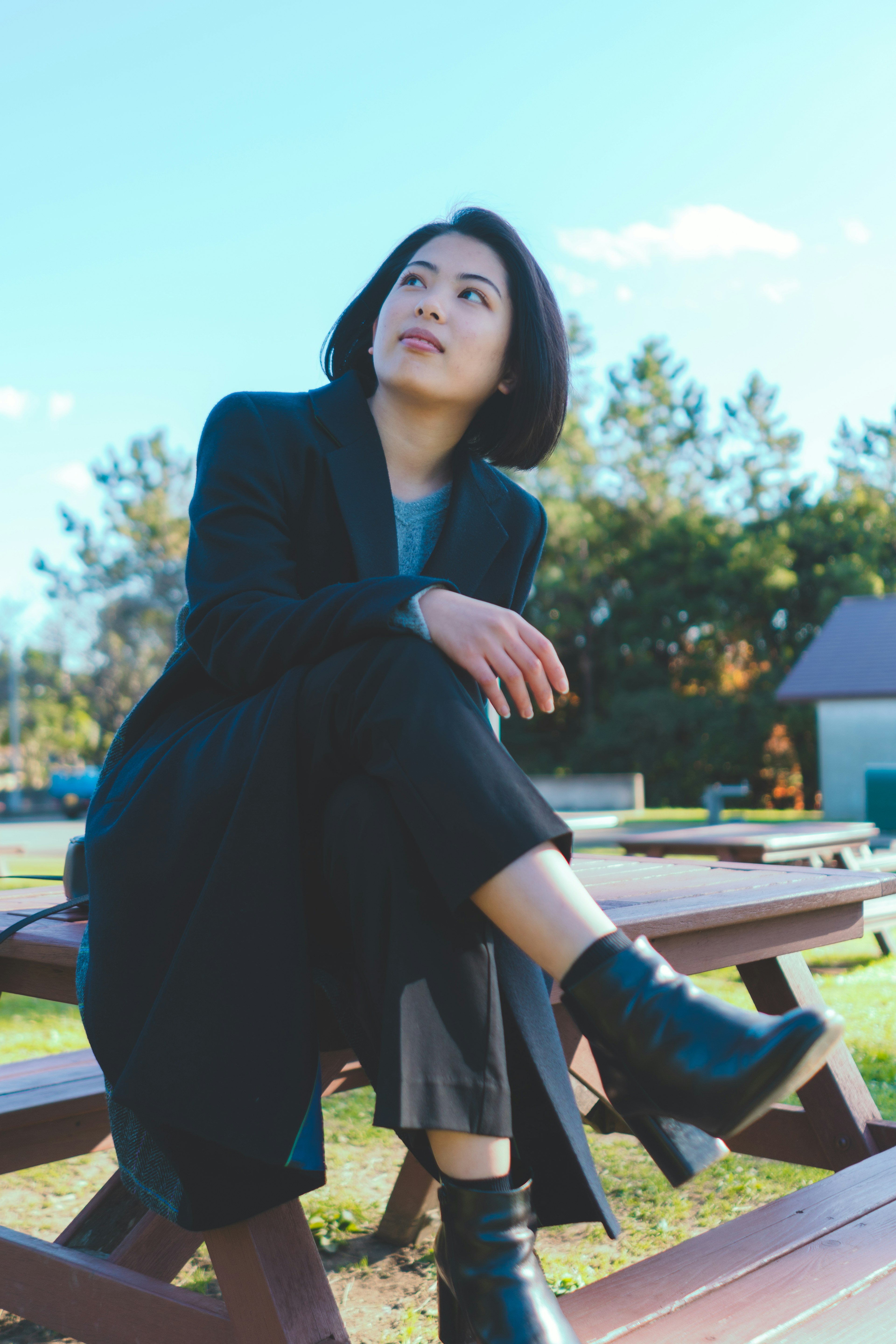 A woman sitting on a park bench wearing a black coat and boots with a blue sky in the background