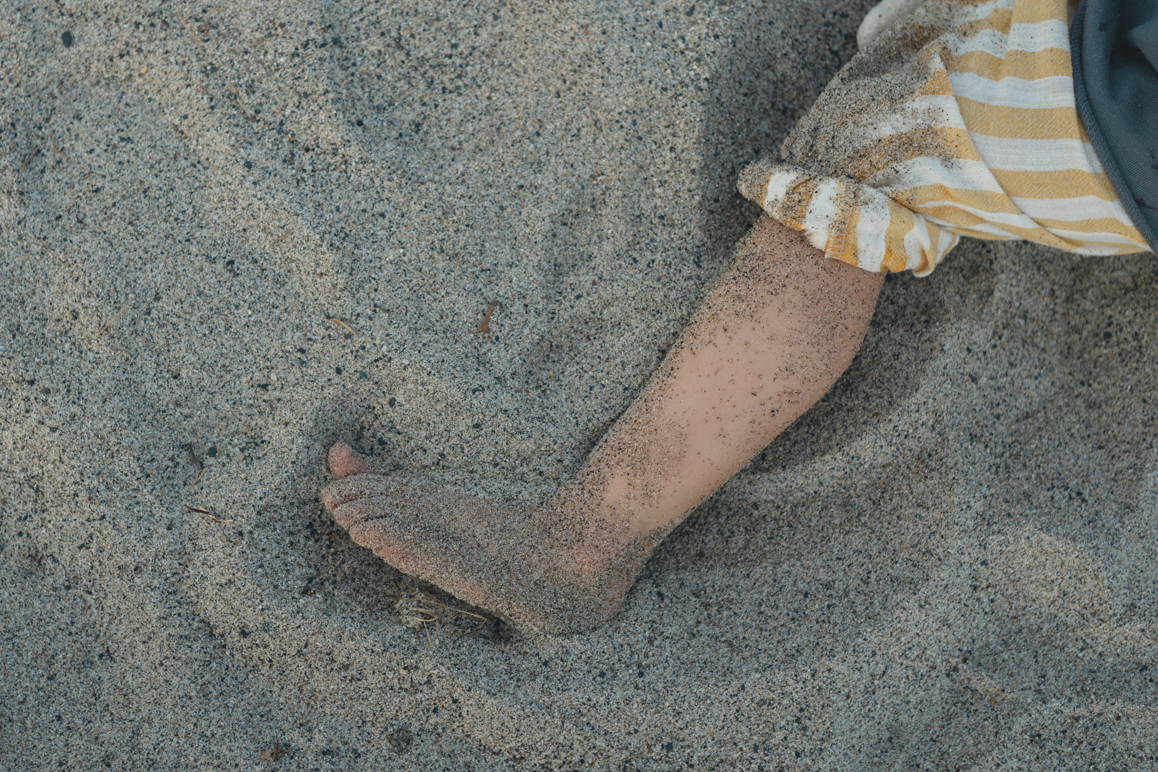 A foot partially buried in sand with a child wearing striped pants