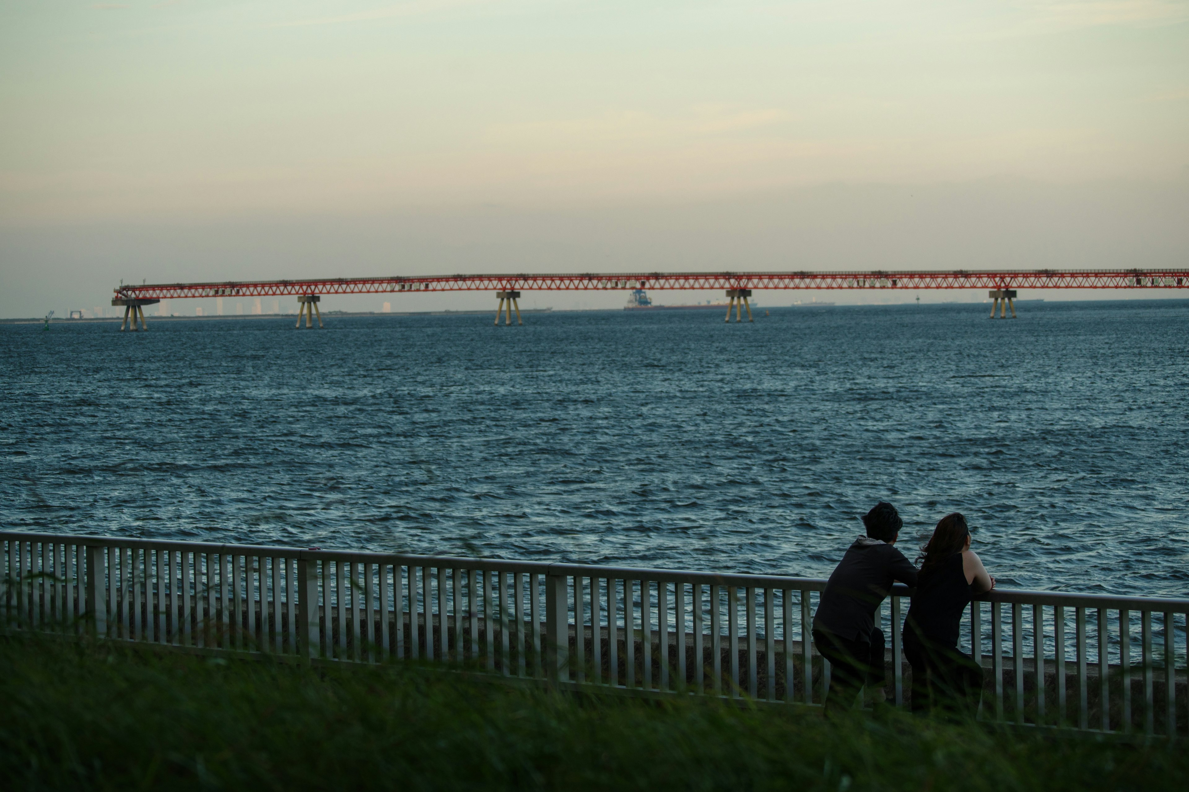 Couple leaning on a fence by the sea with a bridge in the background